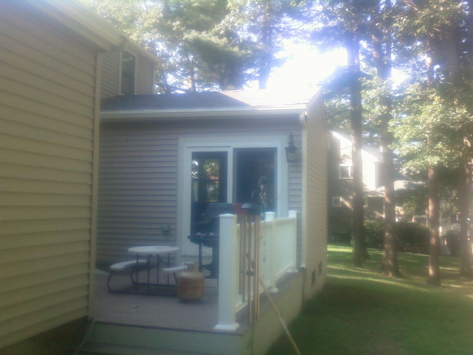 Small deck attached to a house with a sliding glass door. White railing and small round table with chairs. Trees in the background.