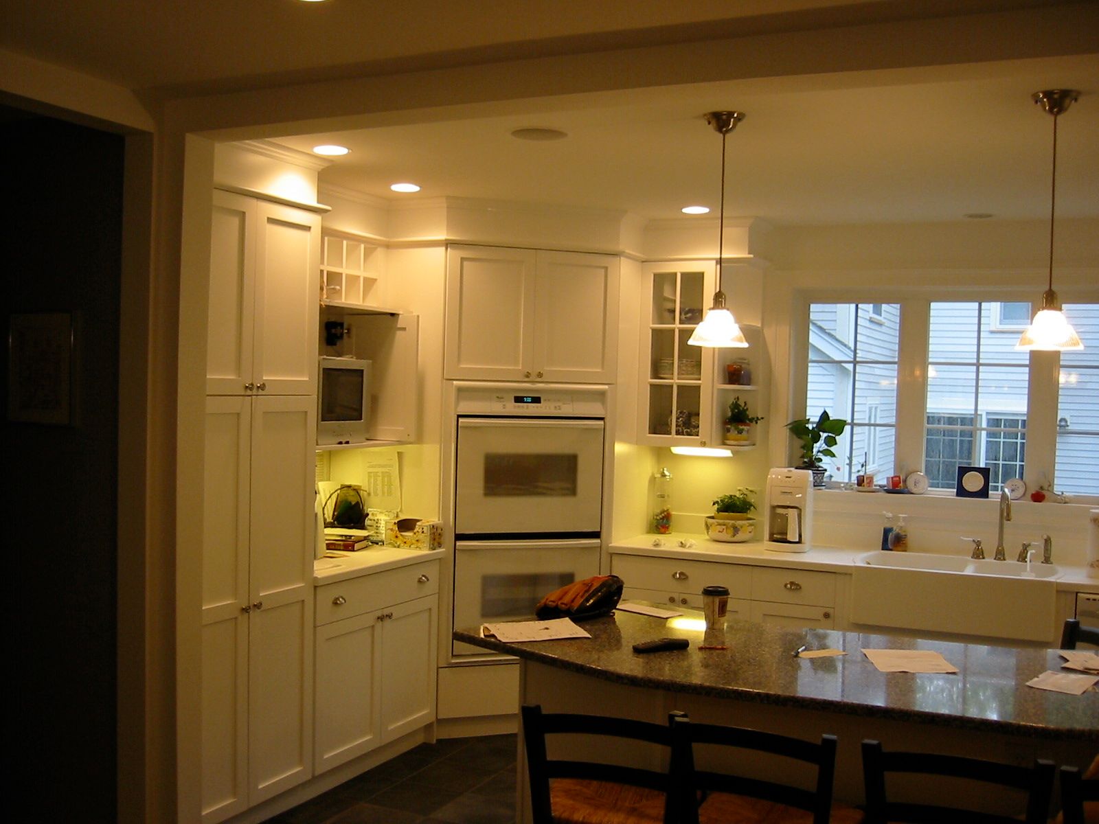 White kitchen with built-in appliances and island with stools. Pendant lights illuminate. Large window in background.