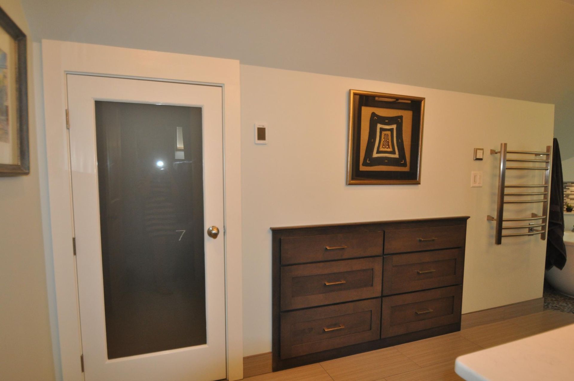 A door, dresser, and art on a beige wall. A towel warmer is on the right.