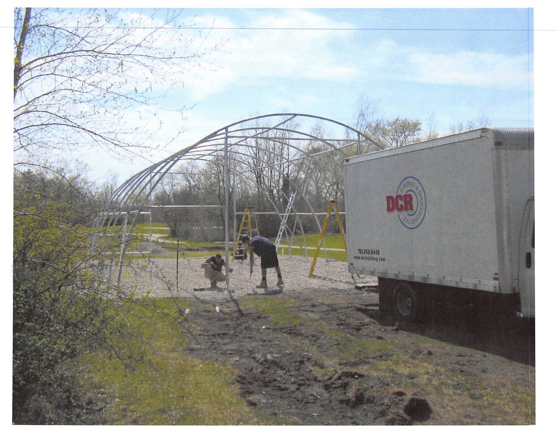 Construction of a large, arched metal structure with workers, next to a white truck on a dirt area.