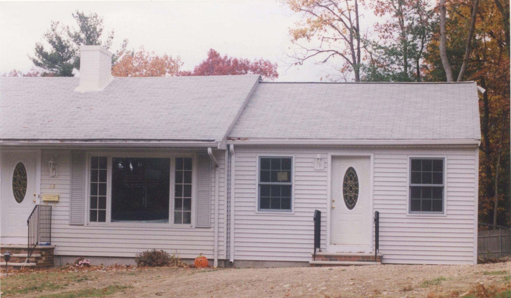 White ranch-style house with a bow window and two doors, set in front of autumn trees.