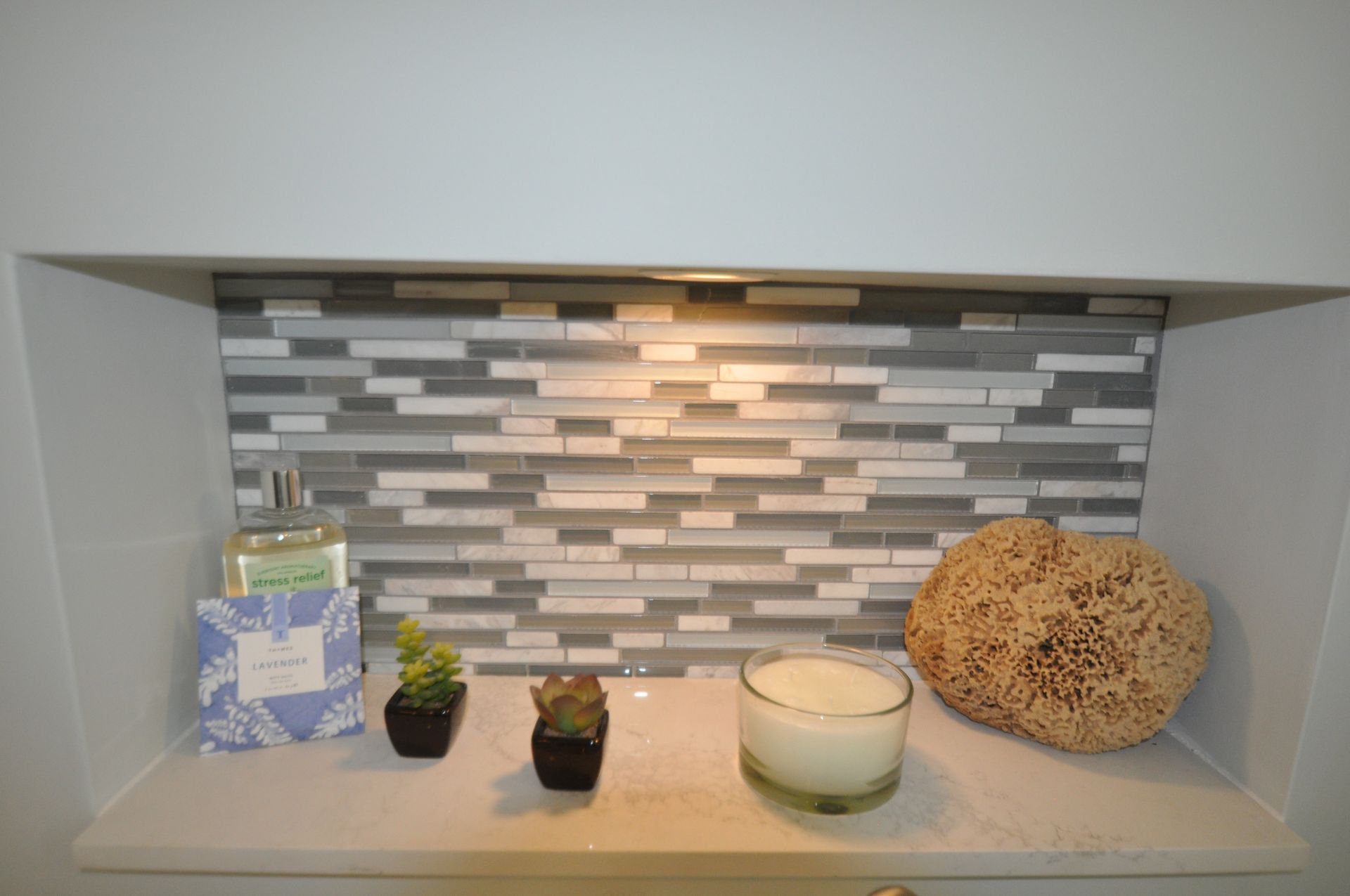 Bathroom niche with toiletries, plants, and a candle. Gray, white, and beige mosaic tile backsplash.