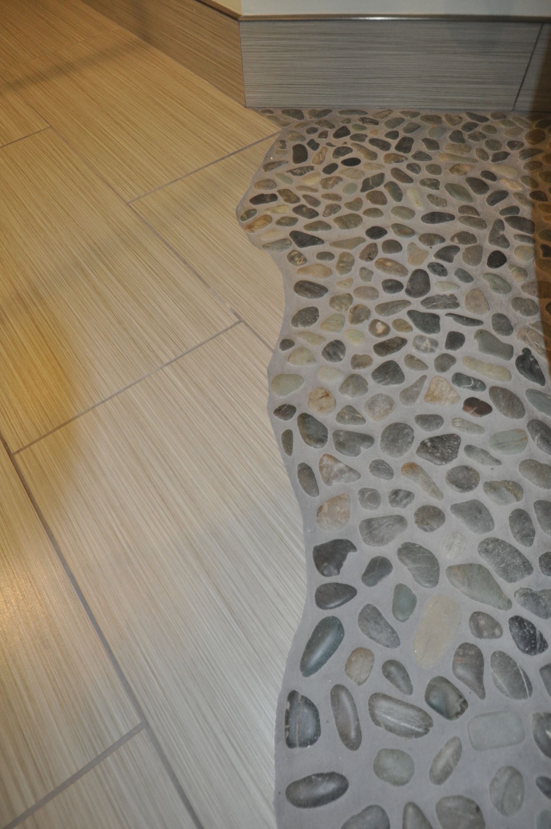 Light brown tile floor meeting a bumpy, stone-covered floor; silver accent trim and corner of a white wall.