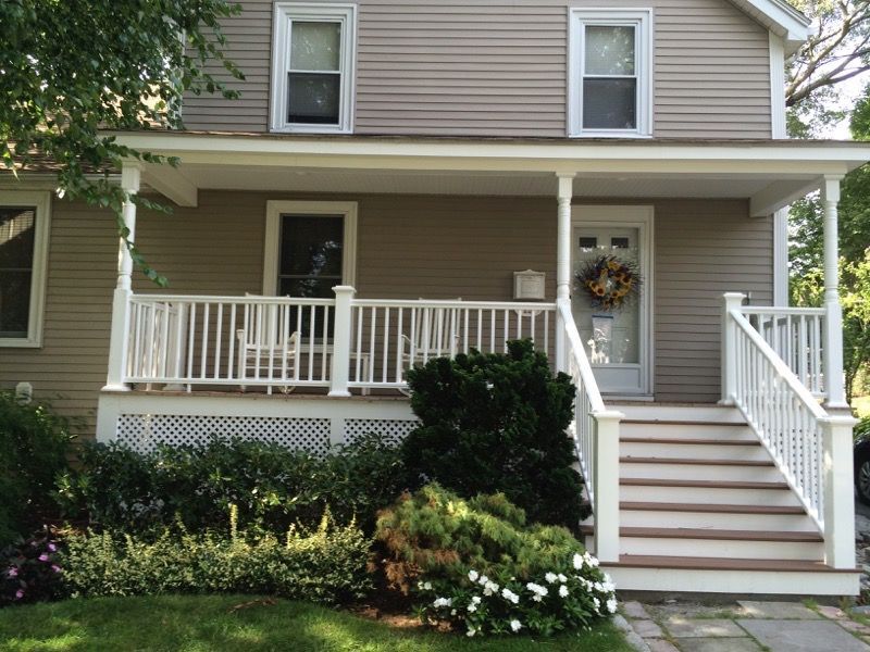 Tan house with white porch, railings, steps, and a wreath on the front door. Landscaping in front.