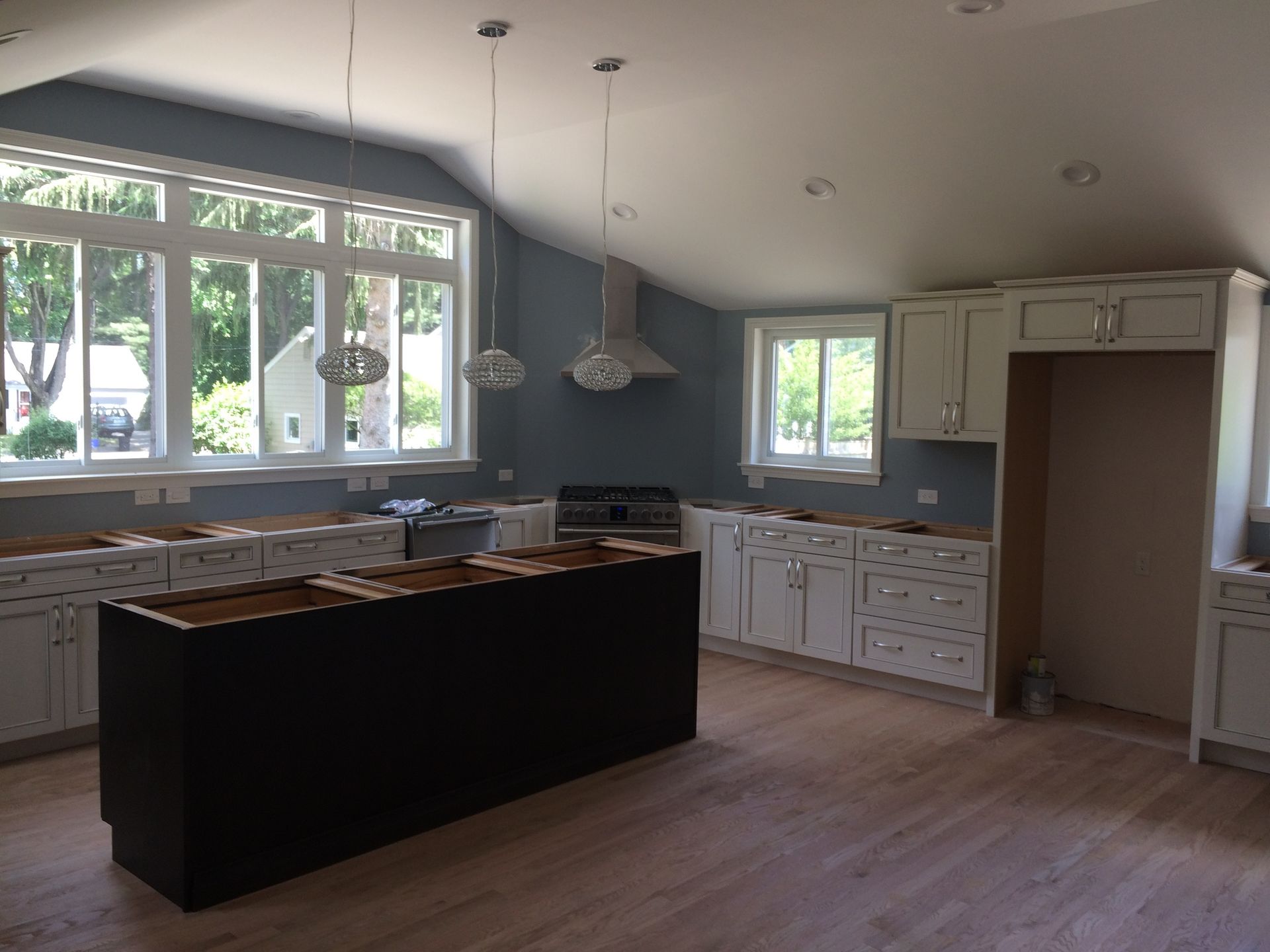 Kitchen with white cabinets, dark island, stainless steel range hood, light blue walls, and large windows.