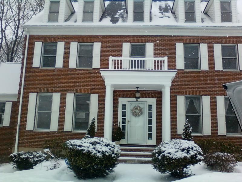 Red brick house with white trim, shutters, and snow-covered bushes in front; snow on roof.