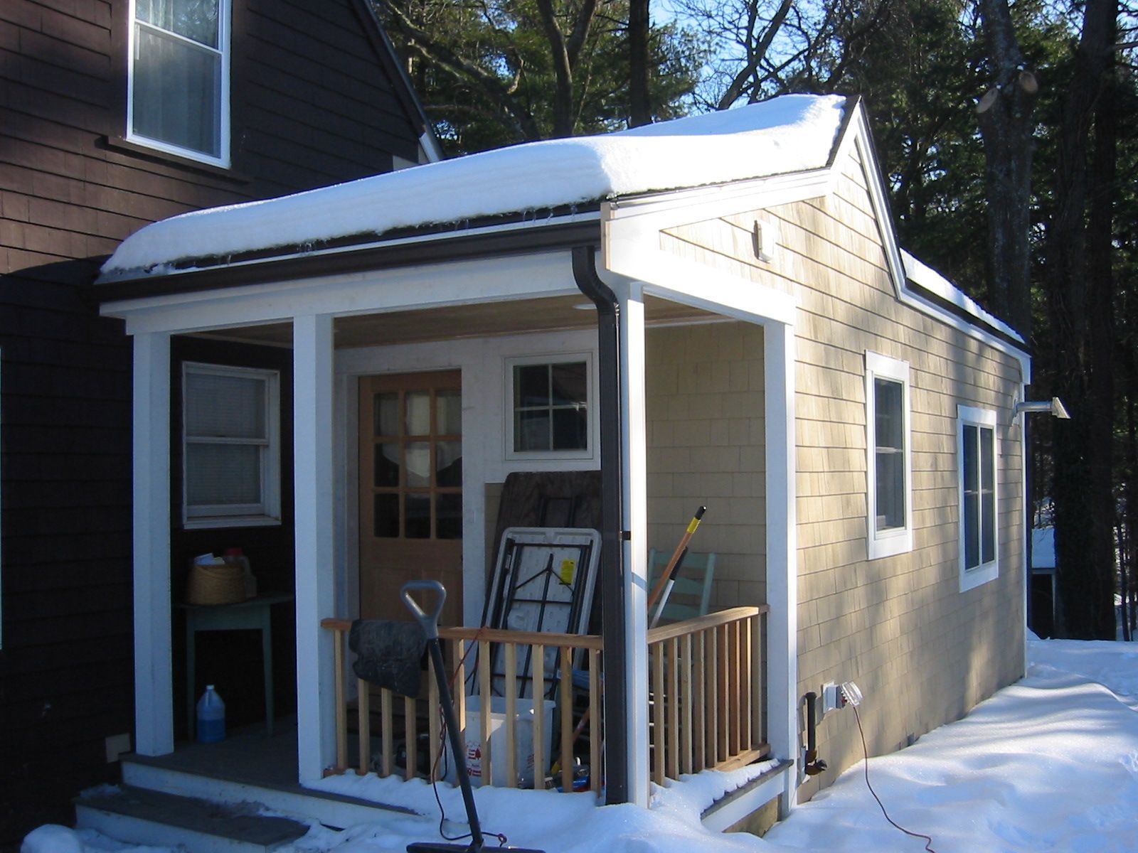 Small tan-colored porch with snow-covered roof, wooden railing, next to dark house in winter.