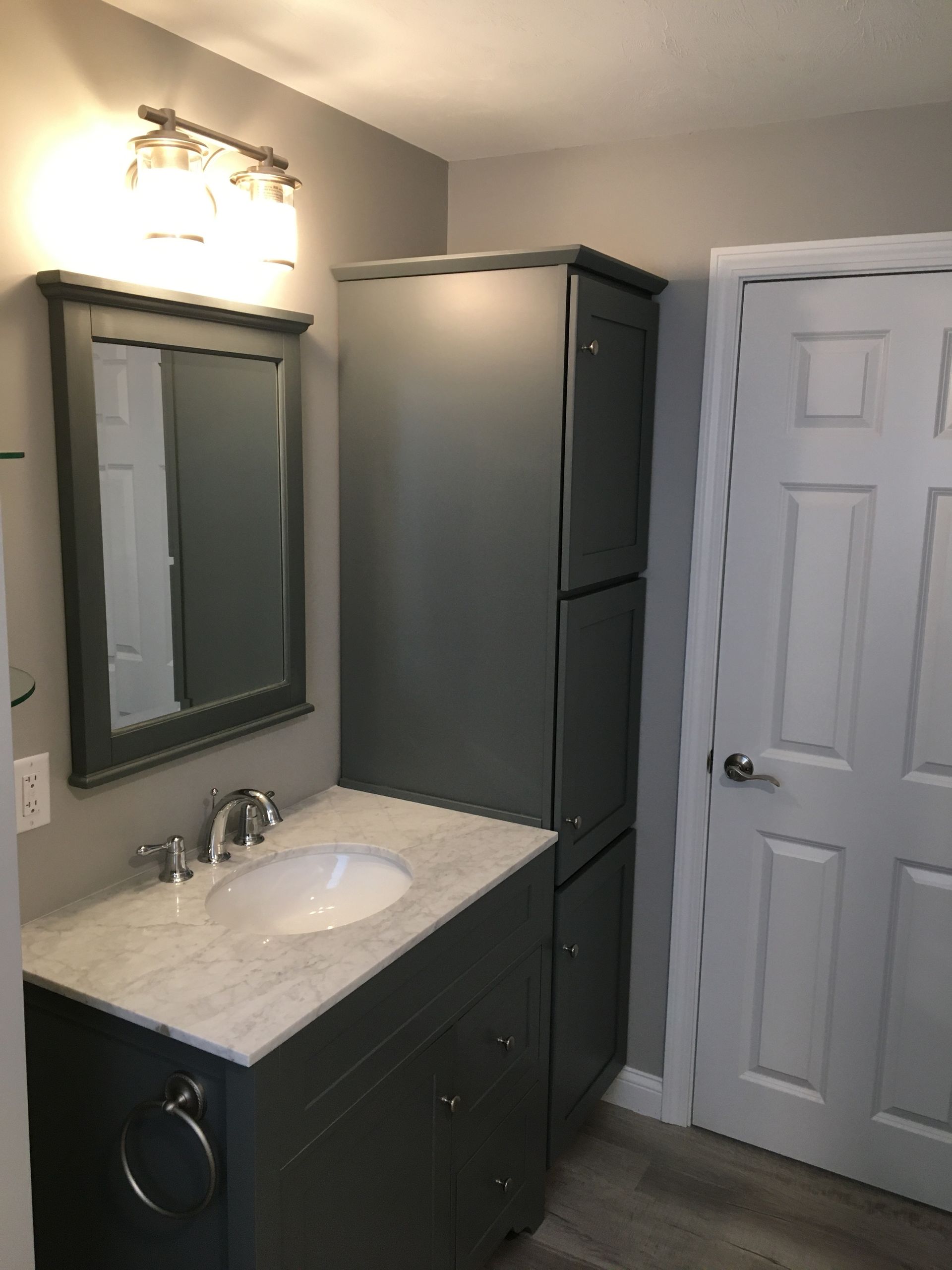 Bathroom with gray vanity, mirror, and tall cabinet; white marble countertop, silver fixtures, and light gray walls.