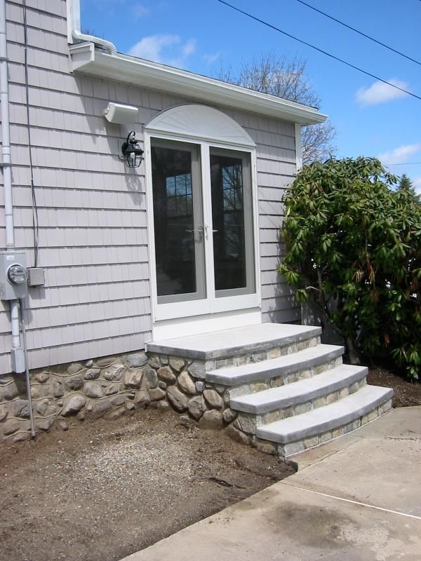 Stone steps leading up to a door, light gray siding, cobblestone foundation, sunny day.
