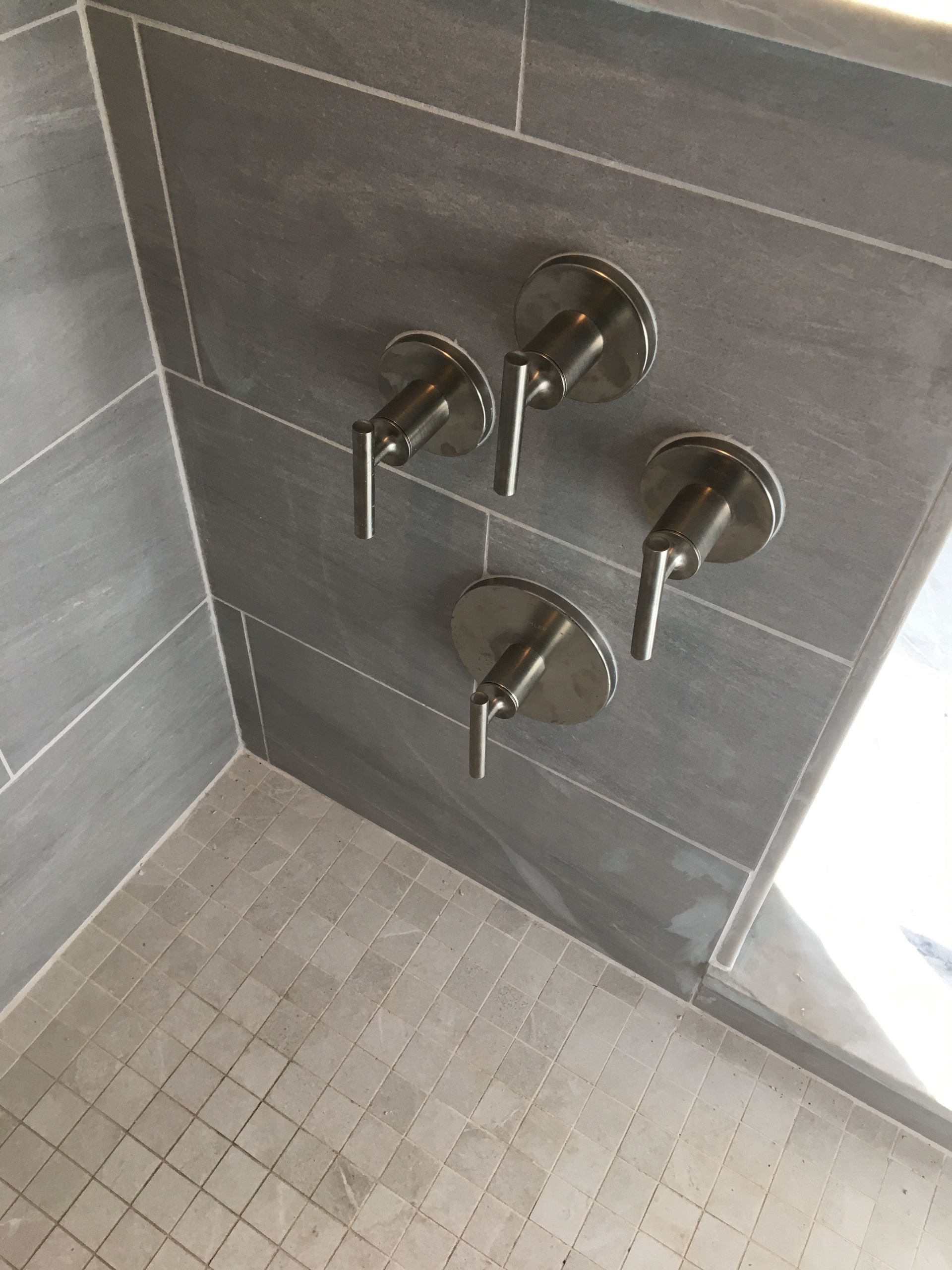 Four silver shower faucet handles on gray tiled wall.