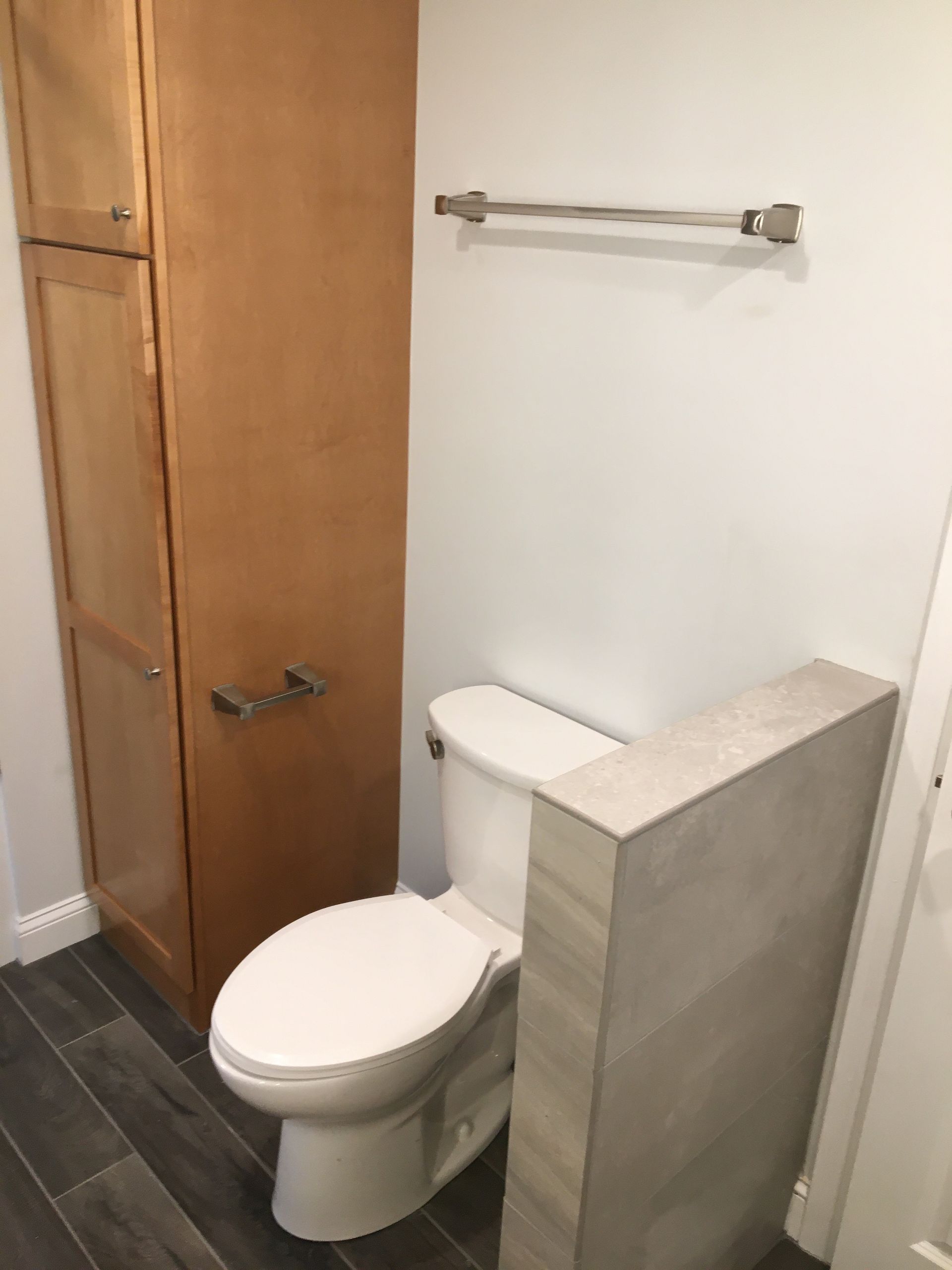 Bathroom with toilet, tall wooden cabinet, towel rack, and tile accents; gray-blue walls.