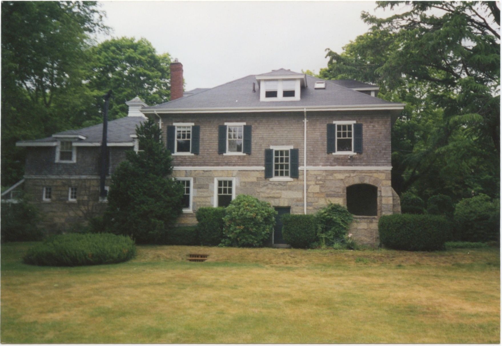 Two-story stone house with gray roof, green shutters, and manicured hedges, viewed from a grassy lawn.