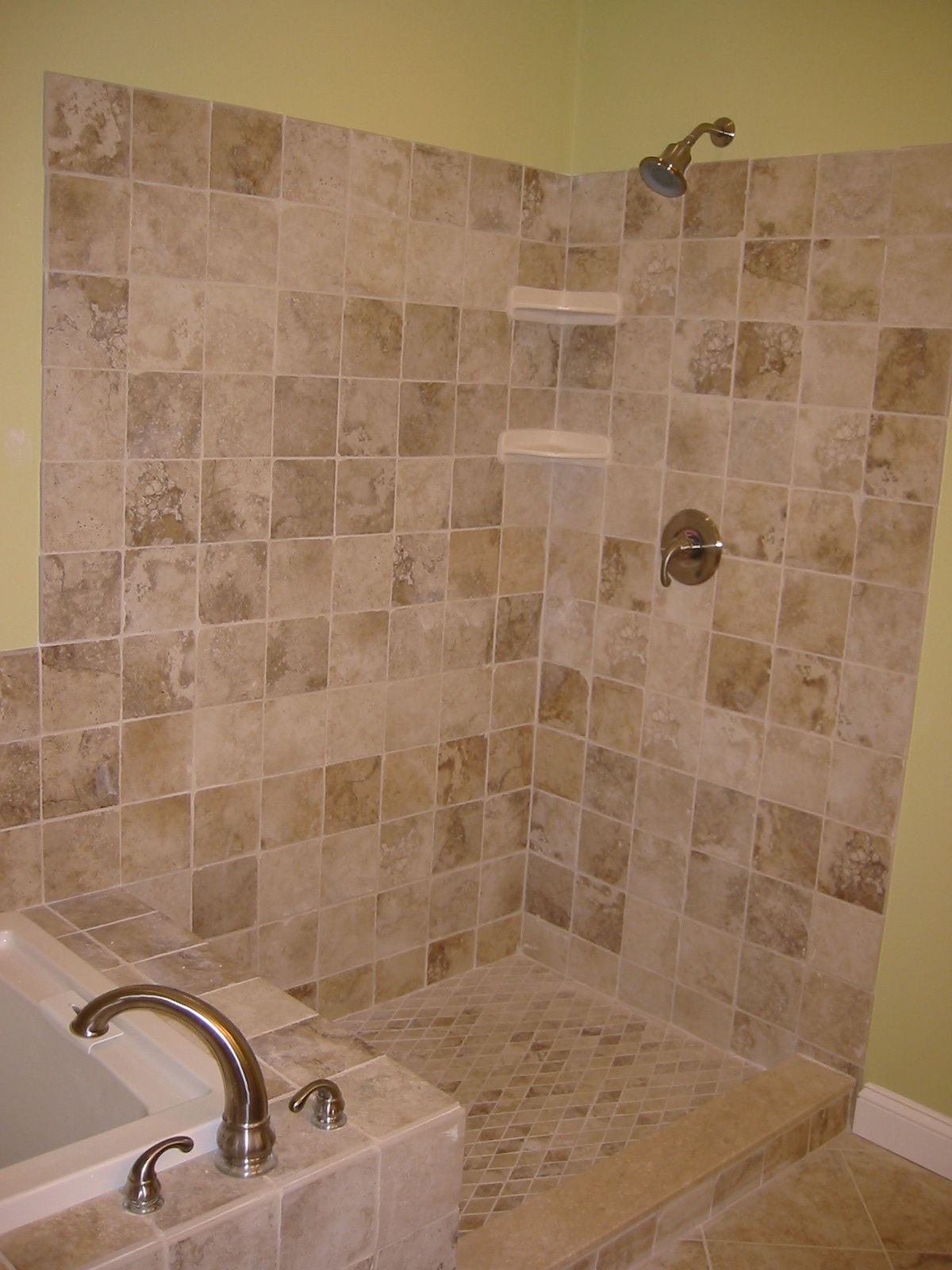 Tiled shower stall with beige and brown square tiles, faucet on the side, and soap shelves.