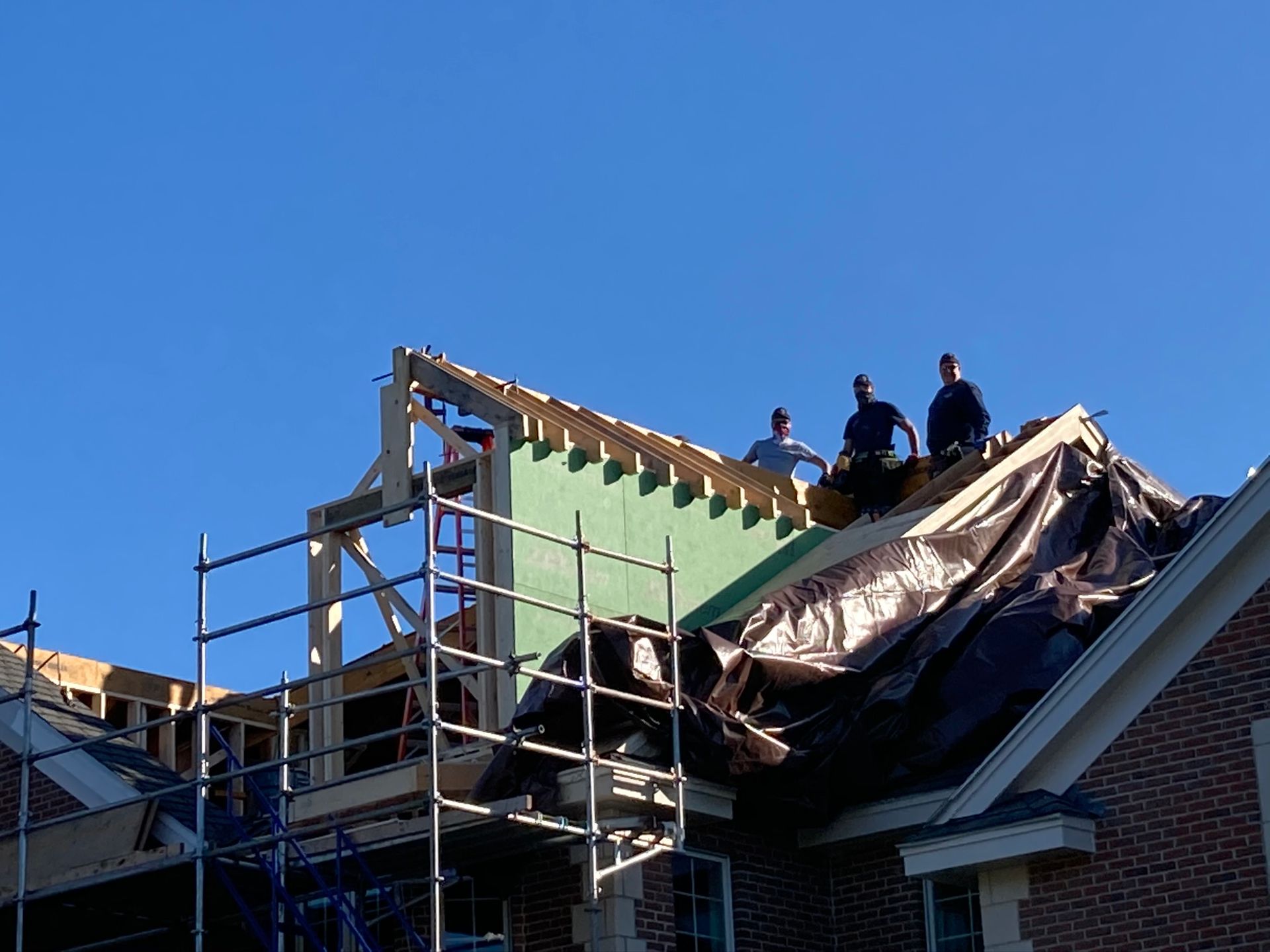 Construction workers on a roof, installing sheathing. Scaffolding, tarp, and blue sky.