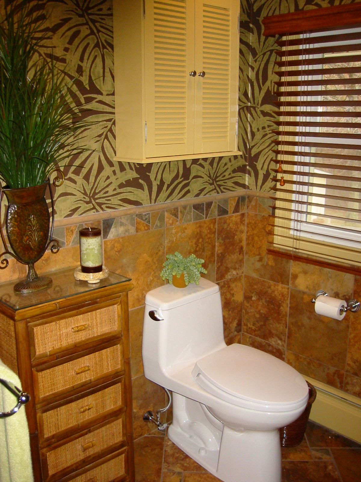 Bathroom with brown tile and floral wallpaper, toilet, cabinet, and window with blinds.