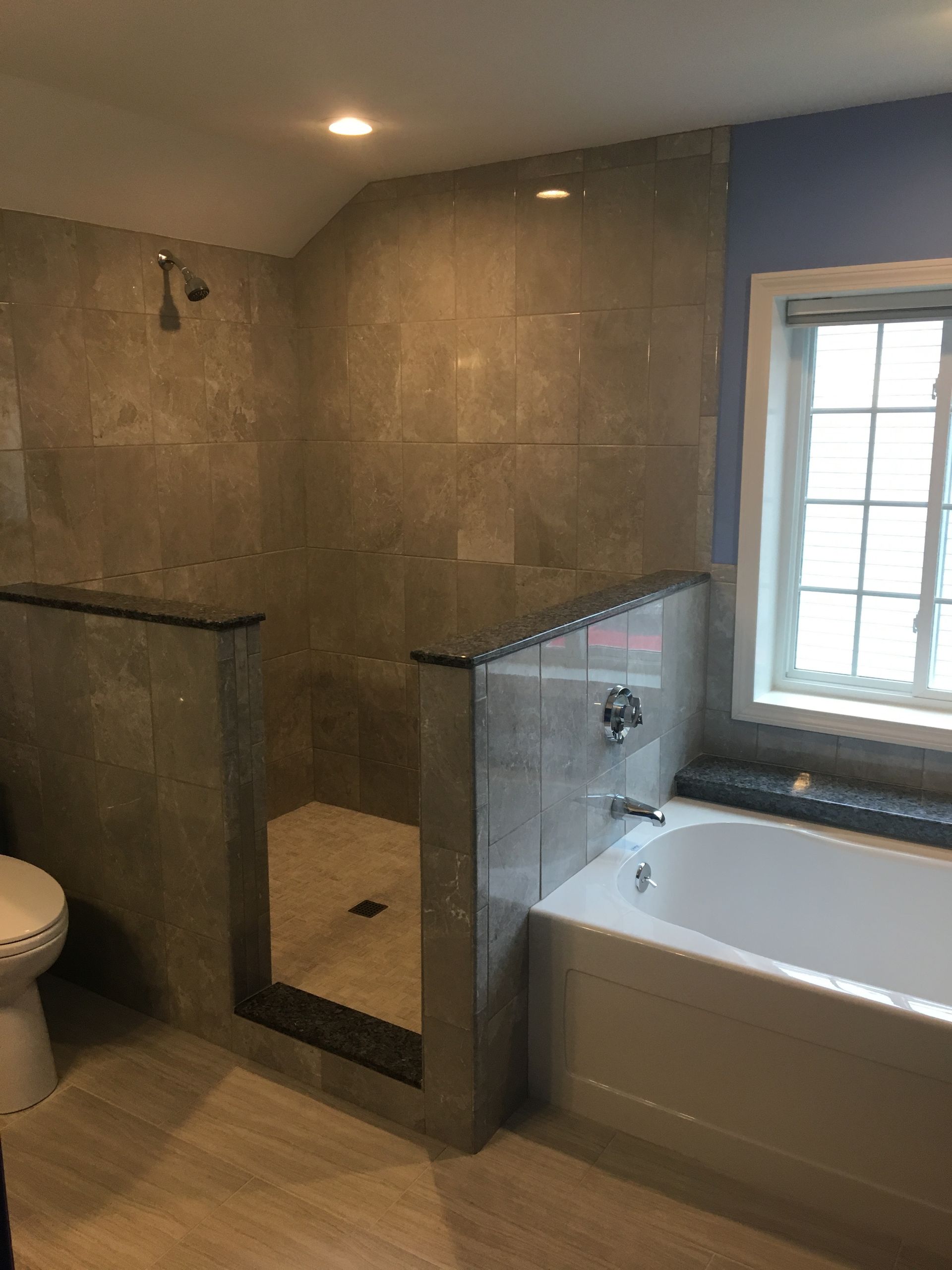 Bathroom with gray tiled shower, white bathtub, and toilet. Blue wall, window, and beige floor.