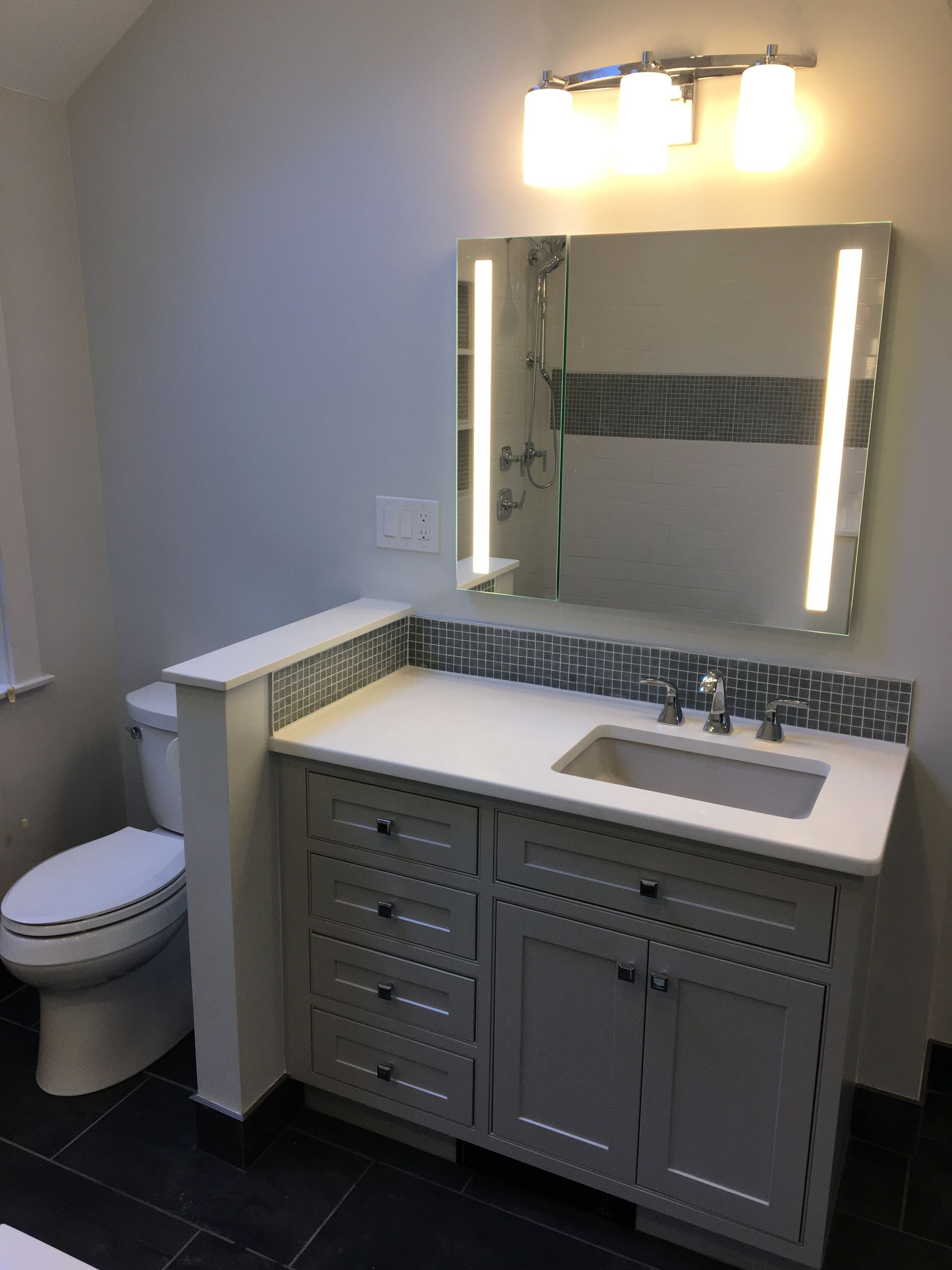 Bathroom with gray vanity, white countertop, blue tile accent, and LED-lit mirror; a toilet is to the left.