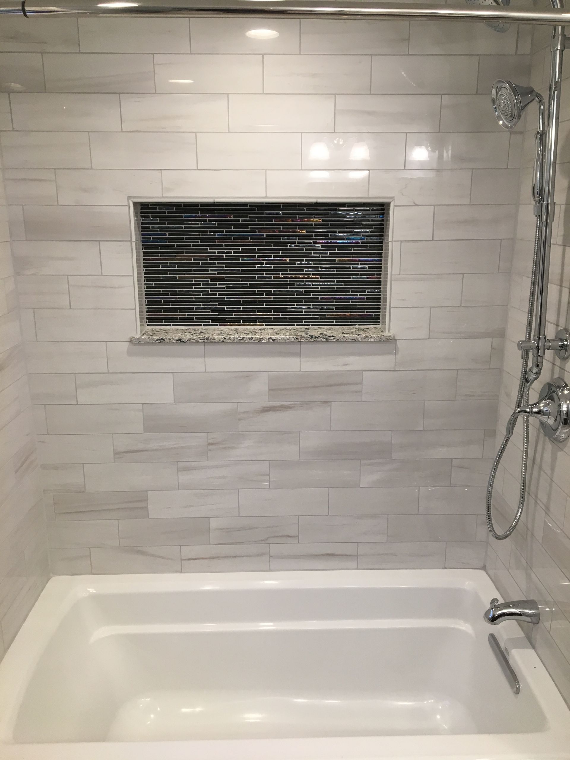 White-tiled bathroom with a recessed shelf of dark mosaic tiles above a white bathtub. Chrome shower fixtures.