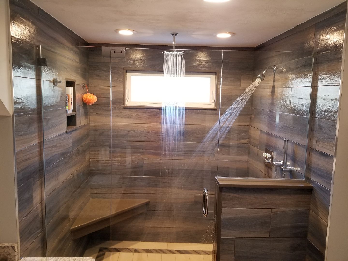 Modern shower with gray-blue wood-look tile, glass doors, bench seat, and water flowing from the overhead showerhead.