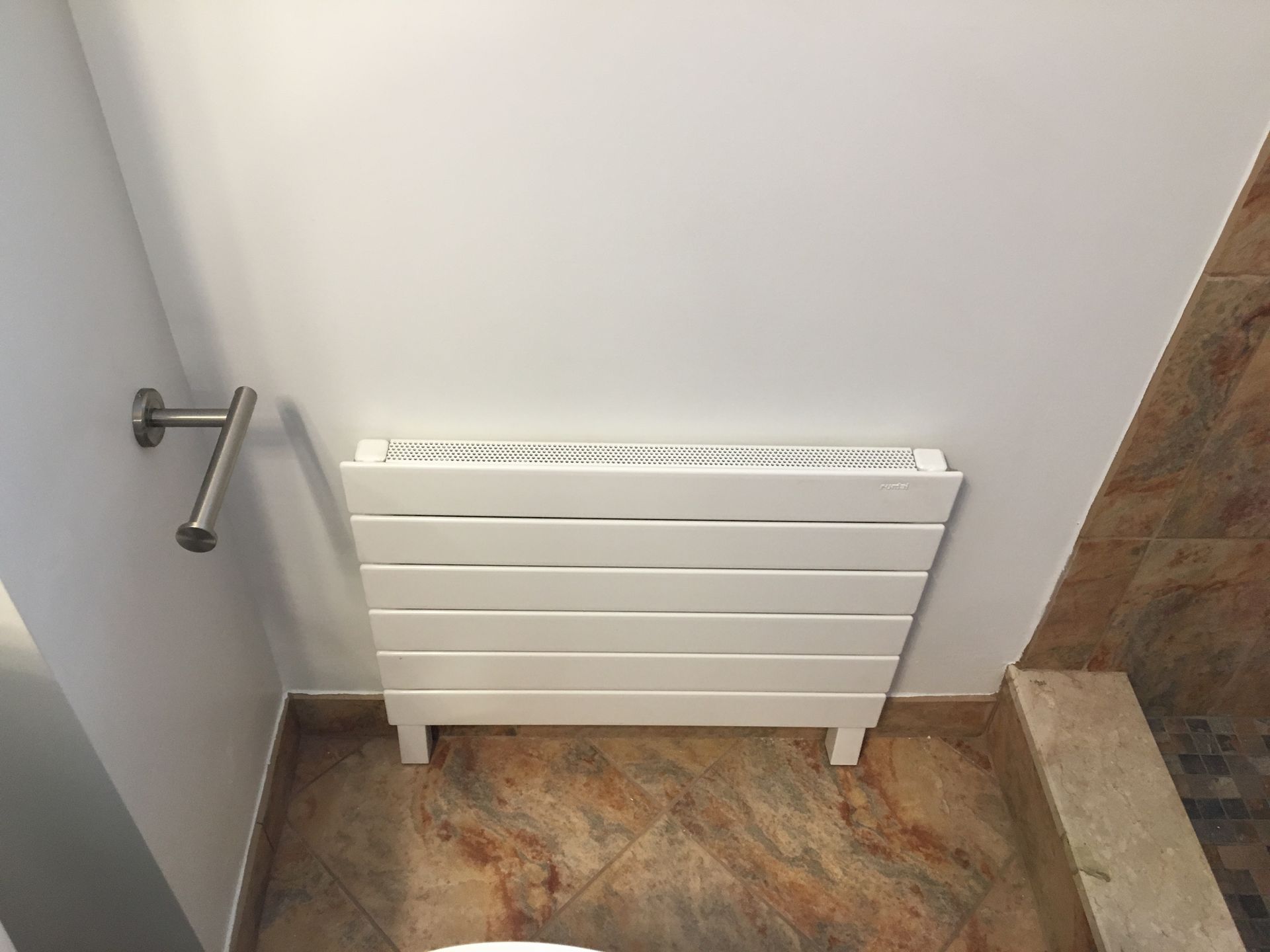 White radiator on a tiled floor, mounted on a white wall next to a towel rack.