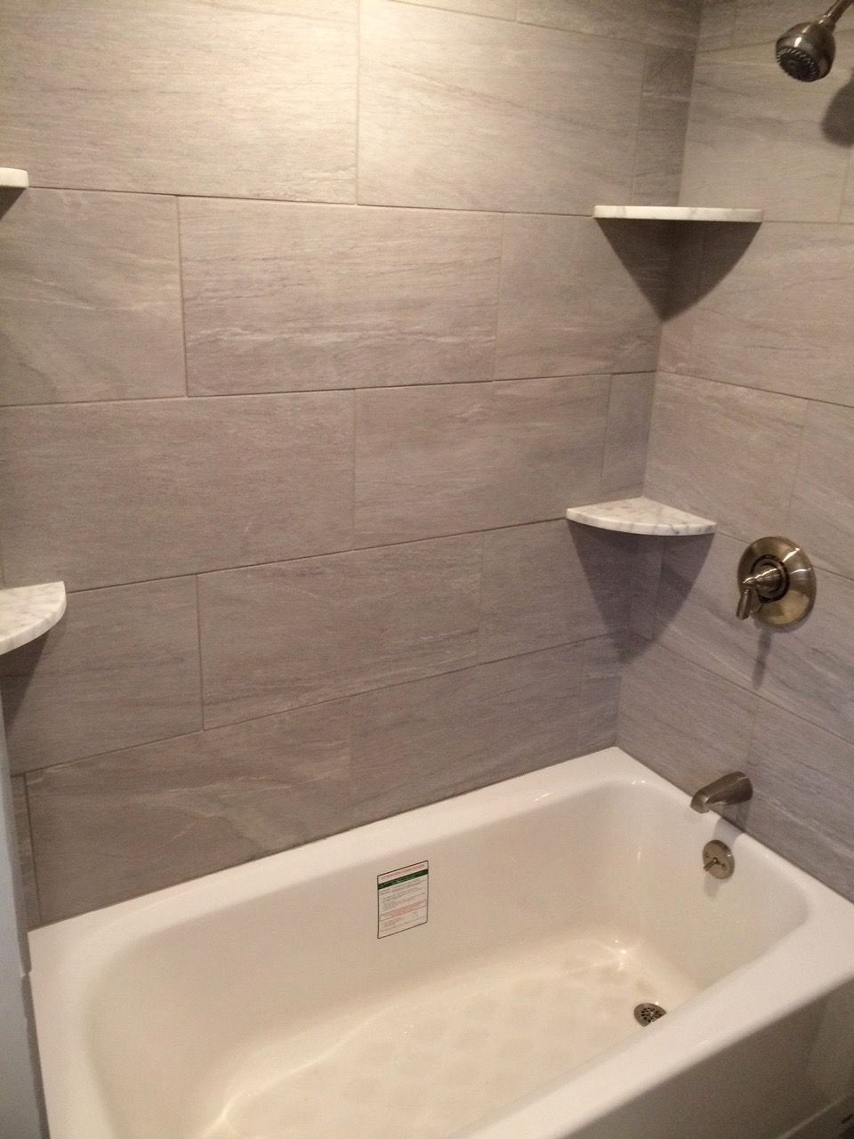 Bathroom with a white bathtub, gray tiled walls, and two white corner shelves.