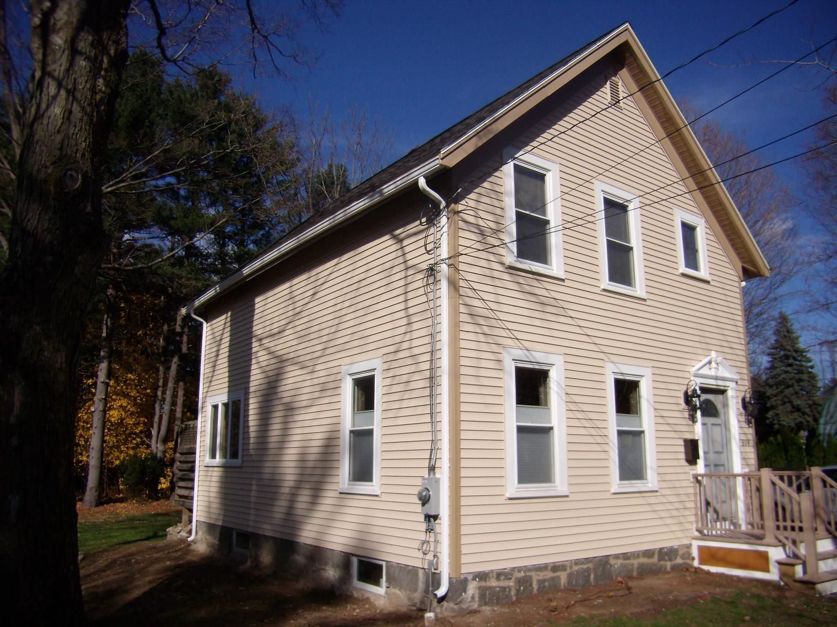Tan two-story house with white trim, multiple windows, and a small front porch, under a blue sky.