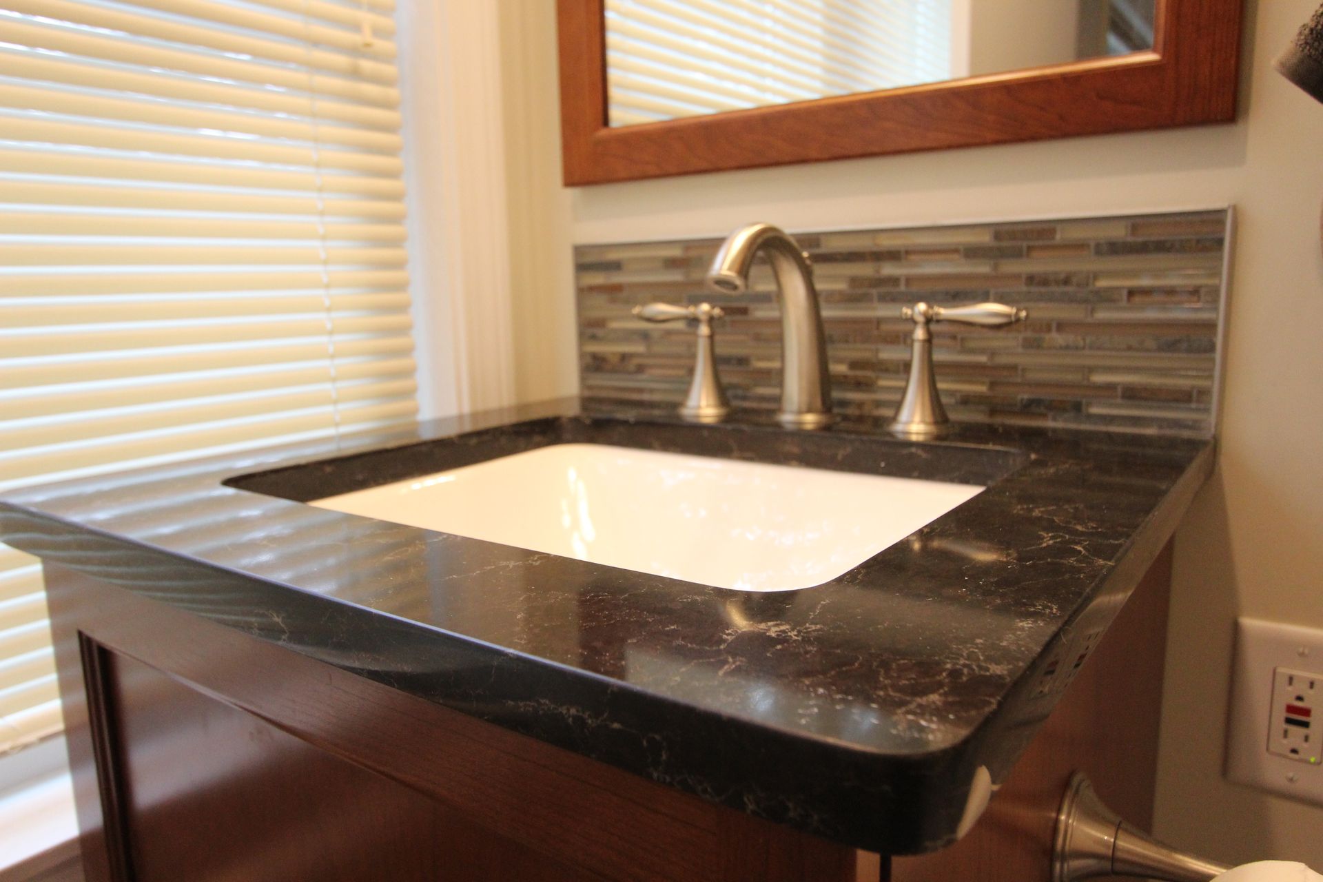 Bathroom sink with dark countertop, backsplash, and blinds.
