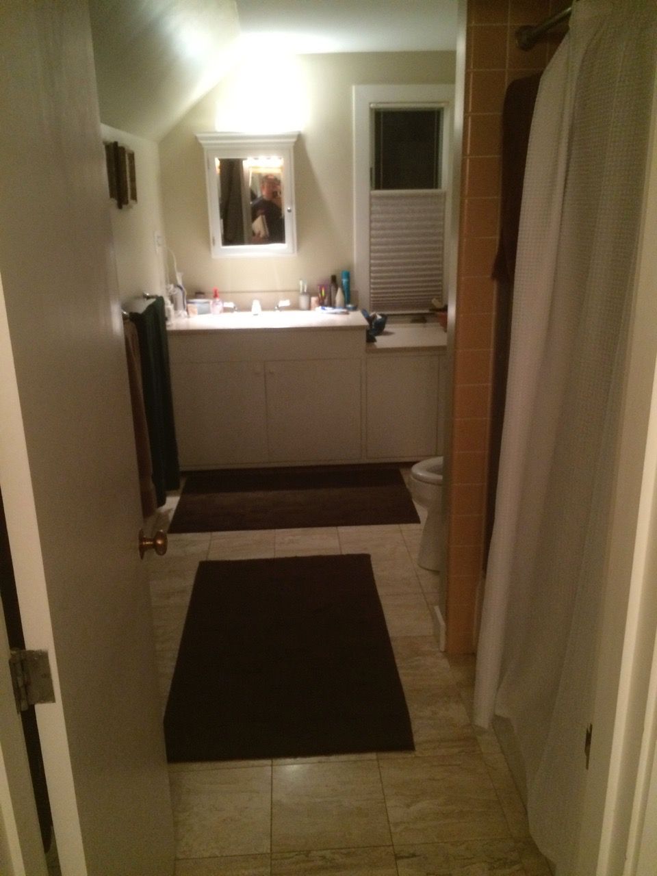 Bathroom: off-white walls, tan tiled floor, brown mats. Vanity with mirror, shower on the right, open door in foreground.