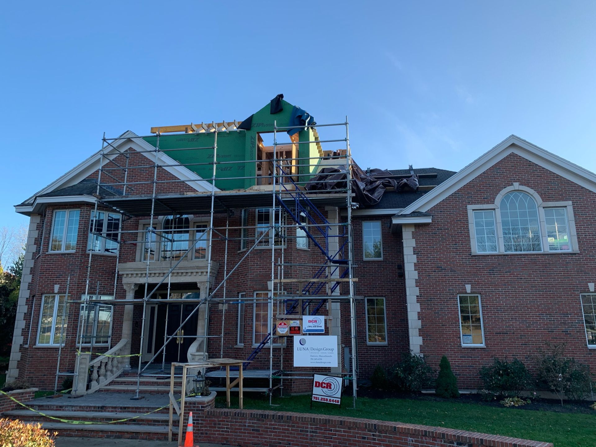 House with exposed roof undergoing repair, scaffolding in front, brick exterior, clear sky.