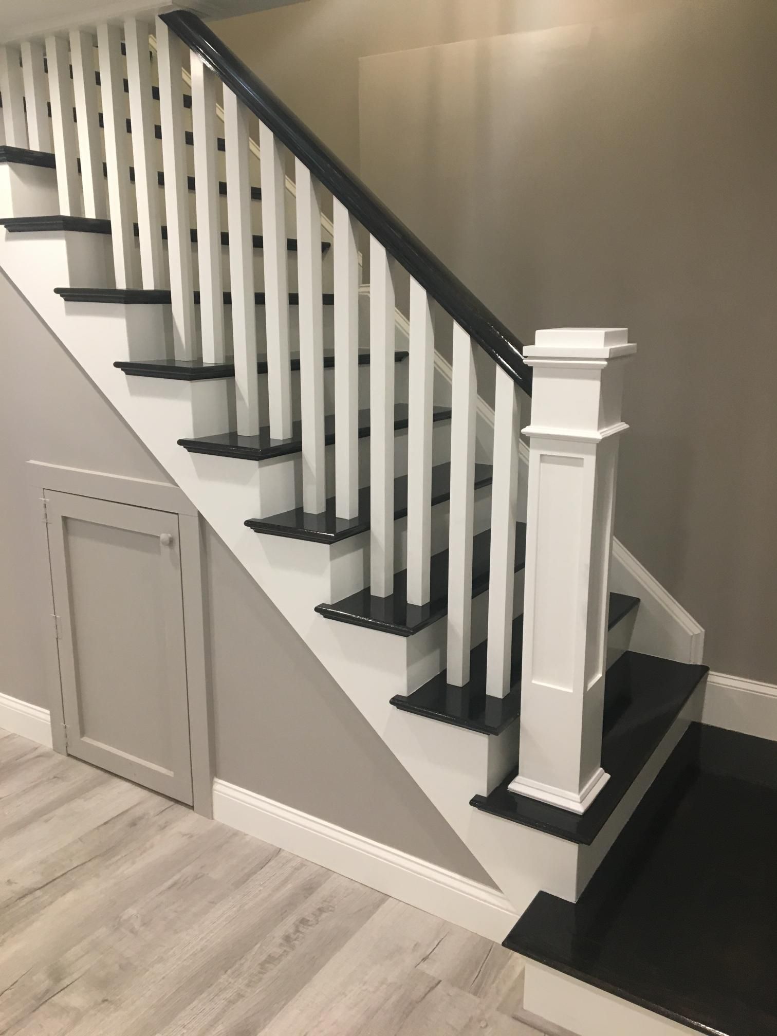 Staircase with white balusters and risers, black steps, and a black handrail. A small door is visible below the stairs.