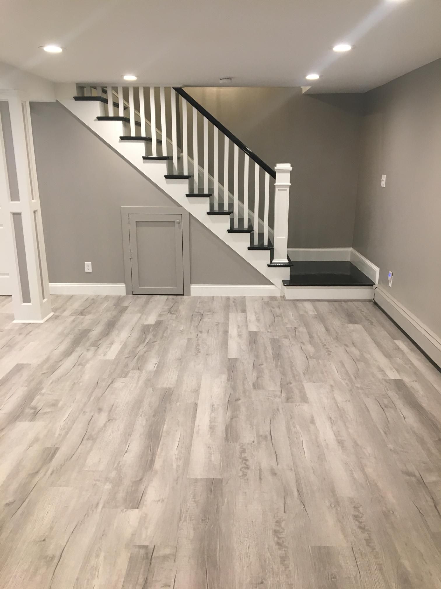 Basement with wood-look flooring, gray walls, white staircase with black handrail, and small gray access door.
