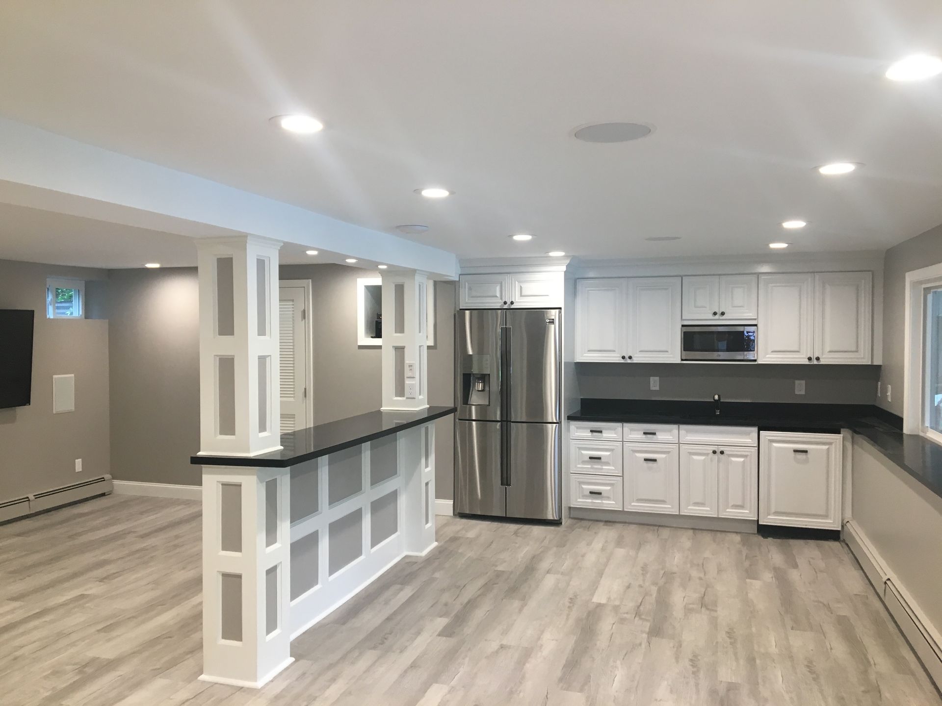 Modern basement with a white kitchen and bar, gray walls, and light wood floors.