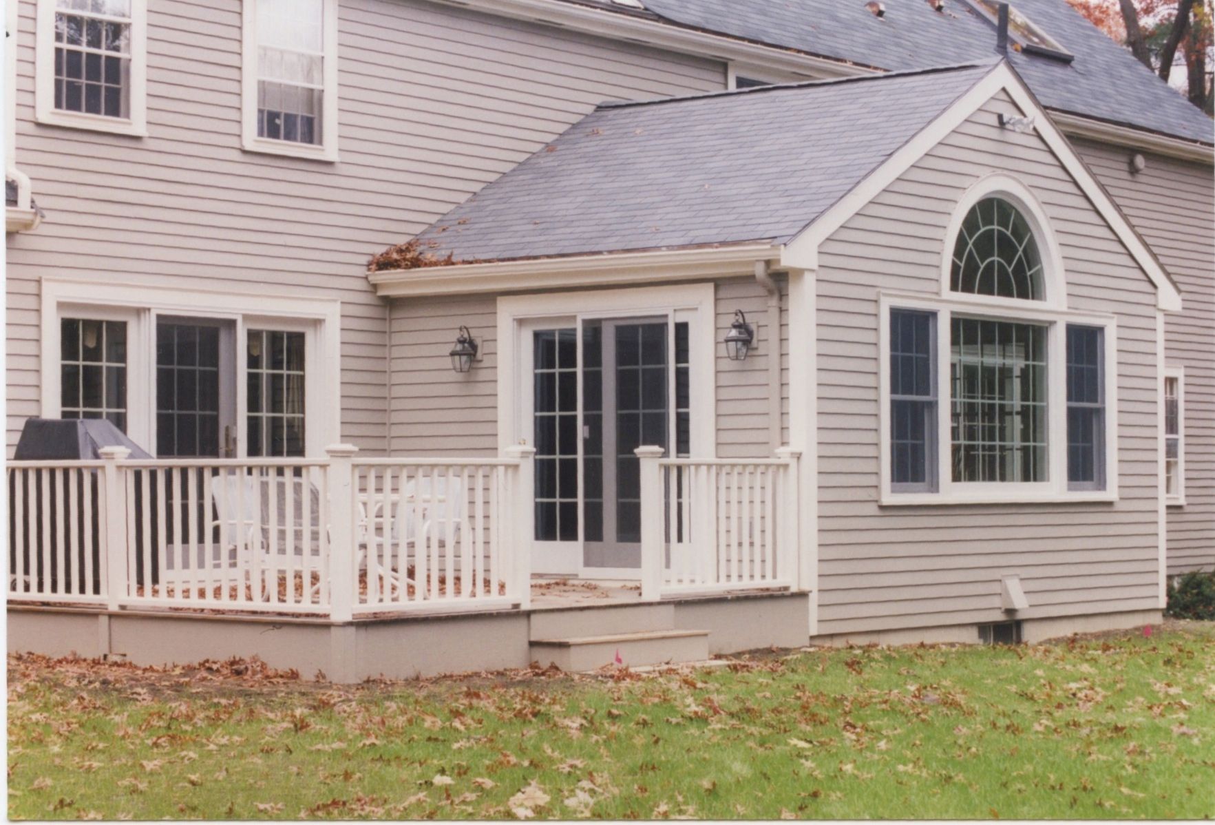Back of a house with a deck and a sunroom. Gray siding, white deck railing, and a dark roof.