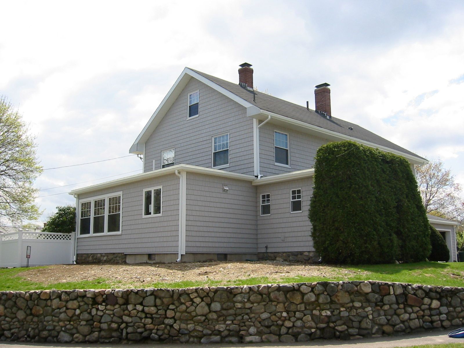 Two-story gray house with a stone wall, green shrubbery, and cloudy sky.