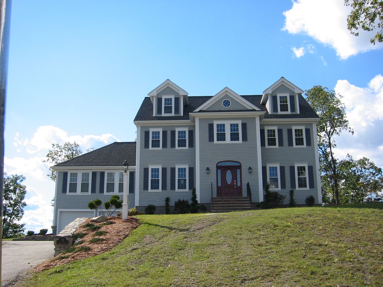 Two-story gray house with blue shutters on a green hillside under a blue sky.