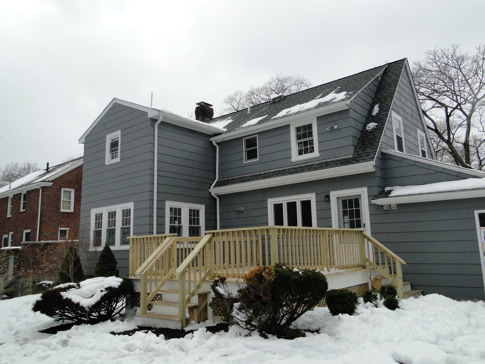 Back of a gray house with a new wooden deck, surrounded by snow.