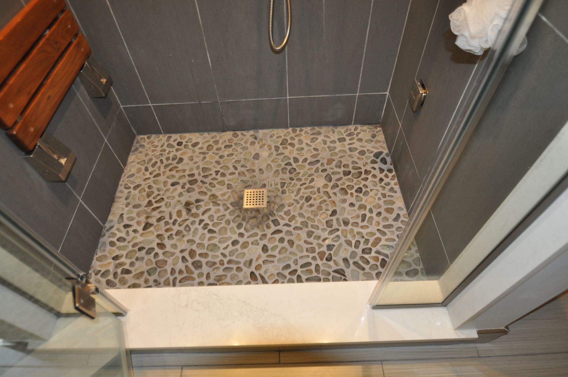 Shower with gray tile walls and a pebble floor; a wooden seat is mounted on the left wall.