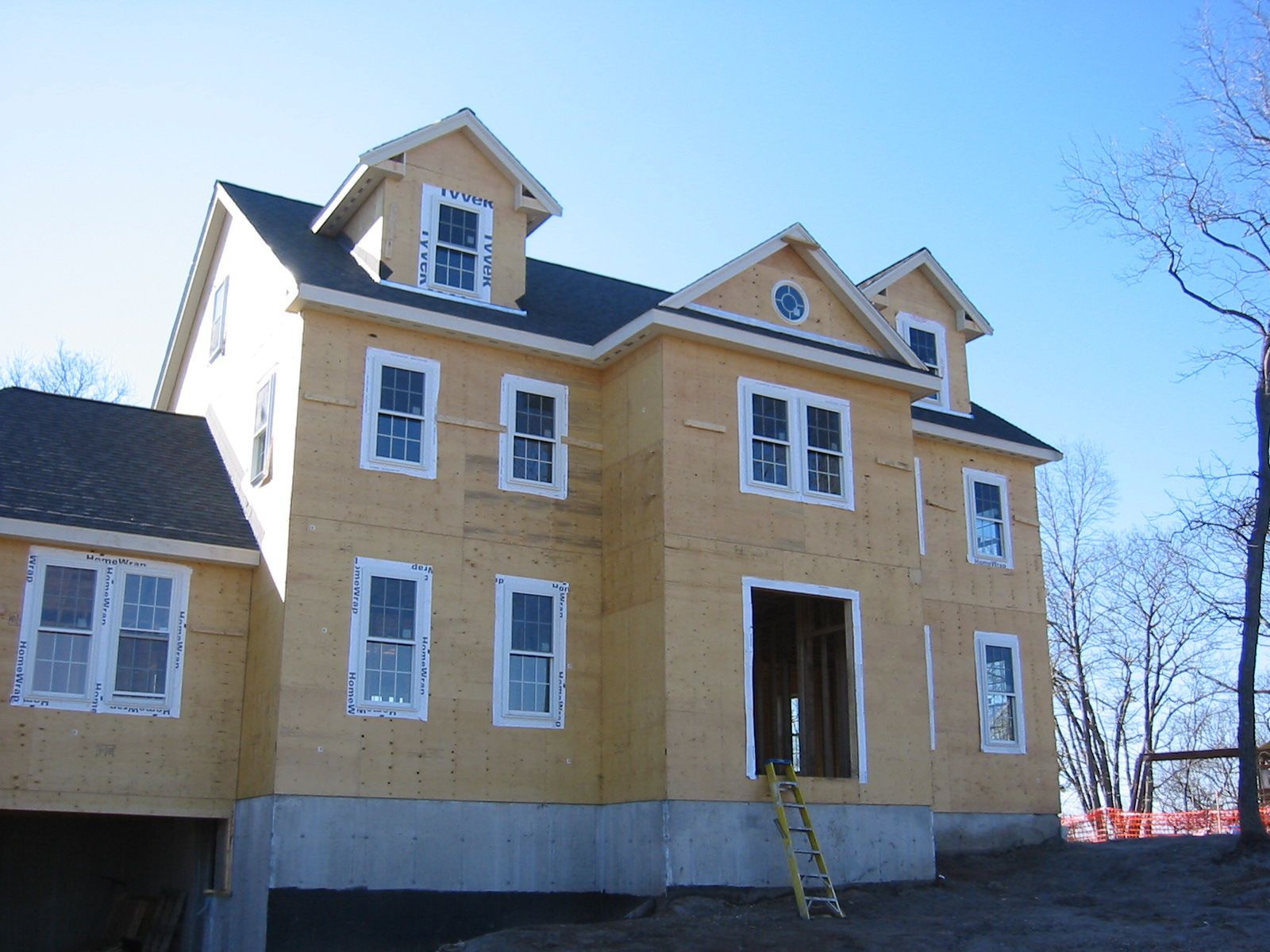 Two-story house under construction with light-colored siding, multiple windows, and dormers under a blue sky.