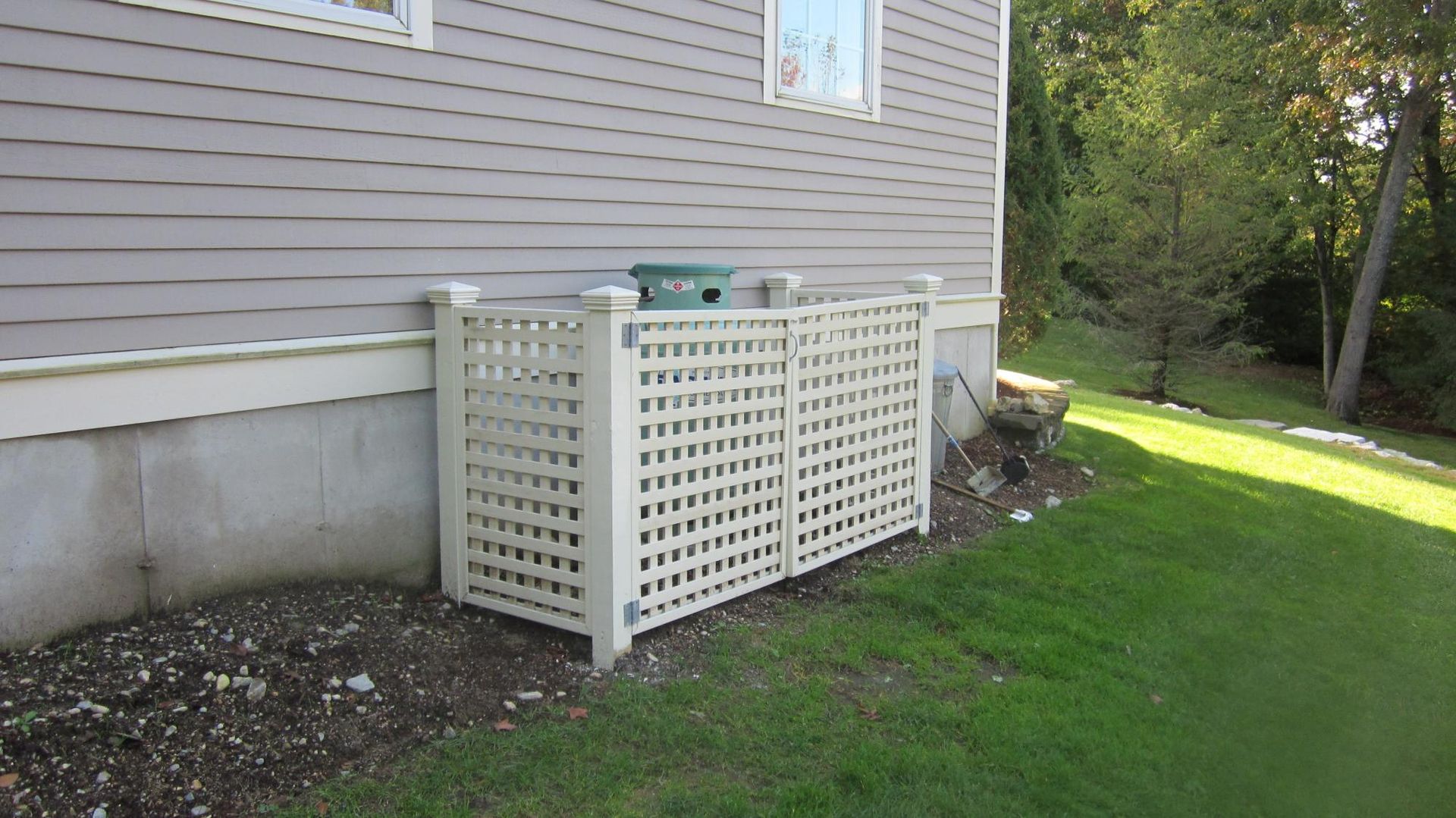 Cream lattice fence encloses an outdoor AC unit next to a house with gray siding on a lawn.