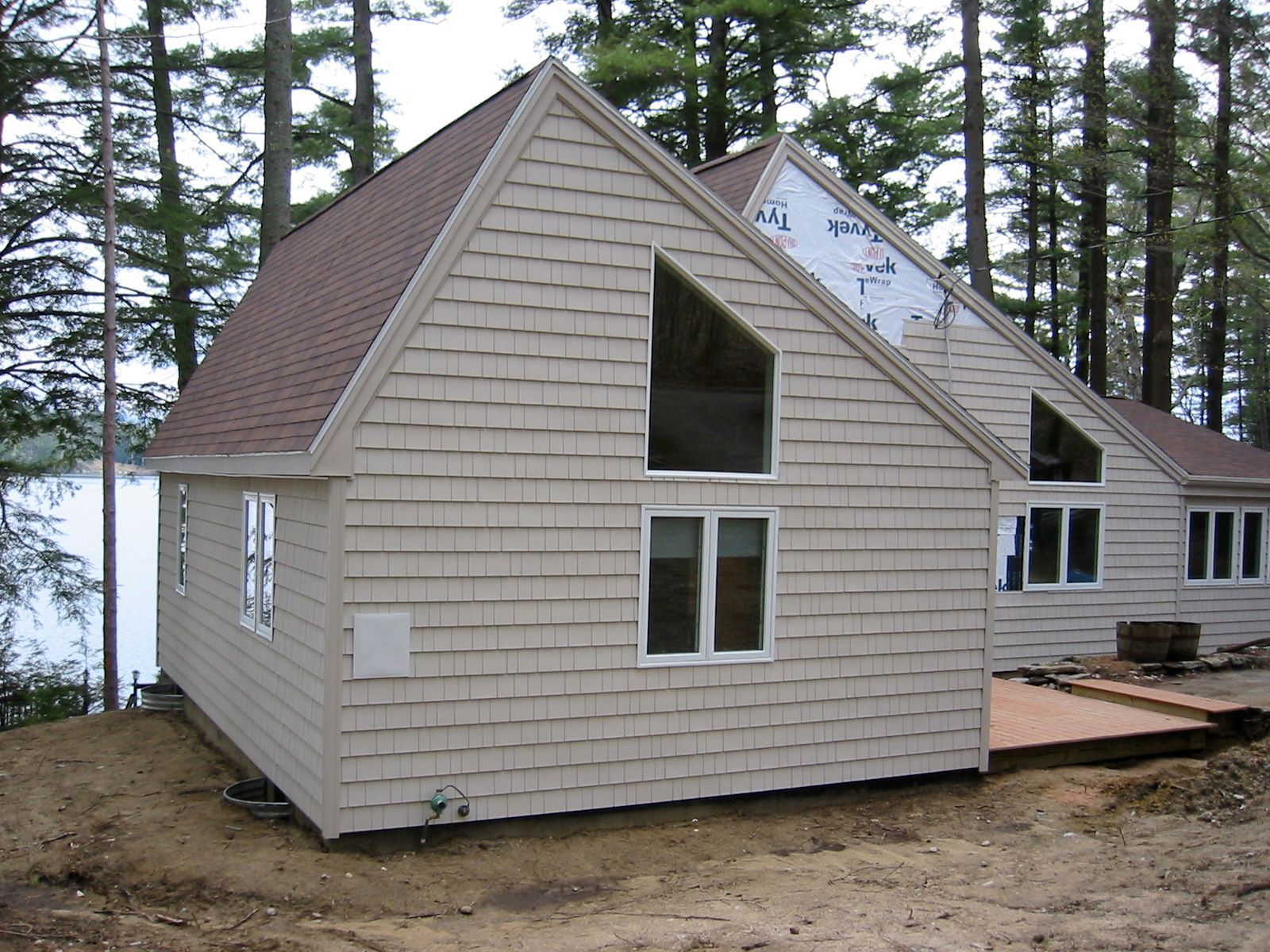 Tan shingle-clad house with a brown roof and triangular windows near a body of water, surrounded by trees.