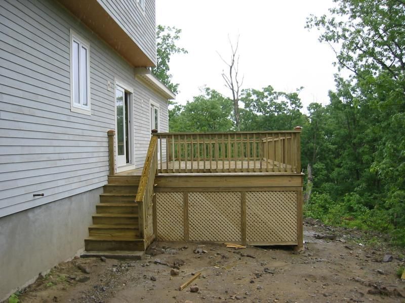 Wooden deck with stairs and lattice skirting attached to a house with gray siding, near a wooded area.