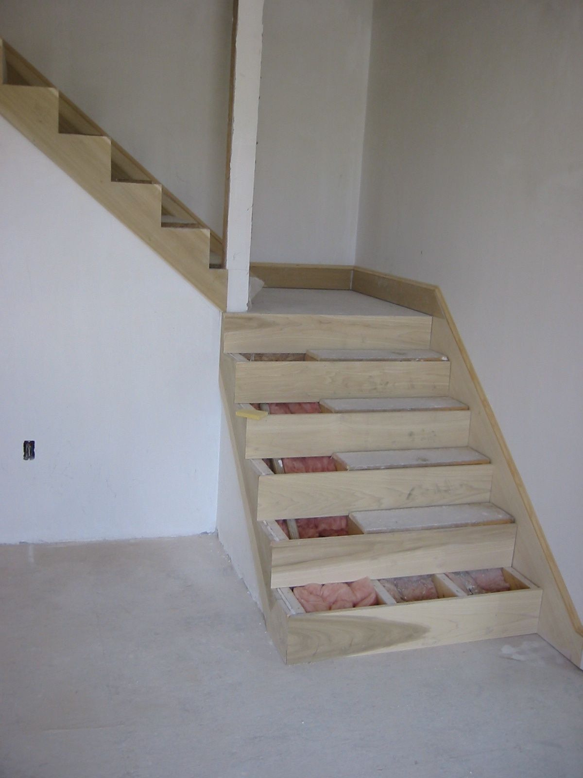 Unfinished wooden staircase with open risers against a white wall in a room under construction.
