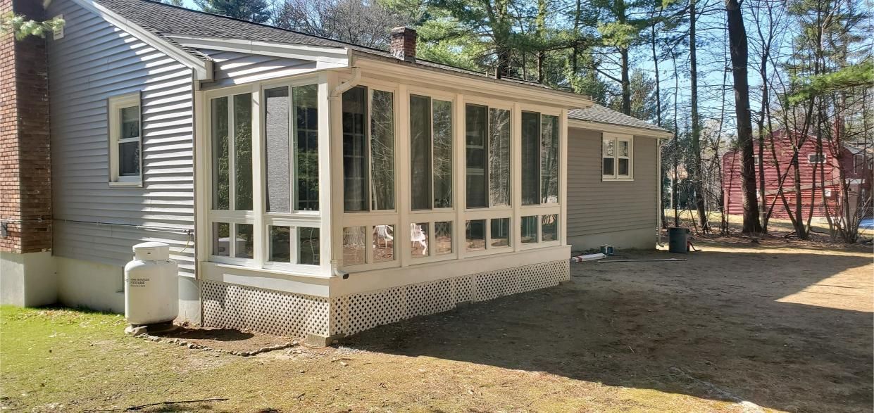 House with screened porch and propane tank on a grassy lawn, surrounded by trees.