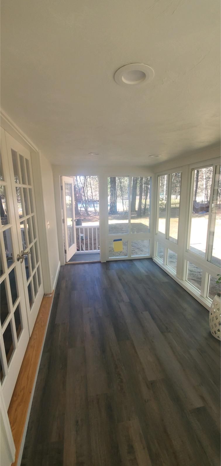 Sunroom with gray wood floors, white trim, and large windows overlooking a wooded area.