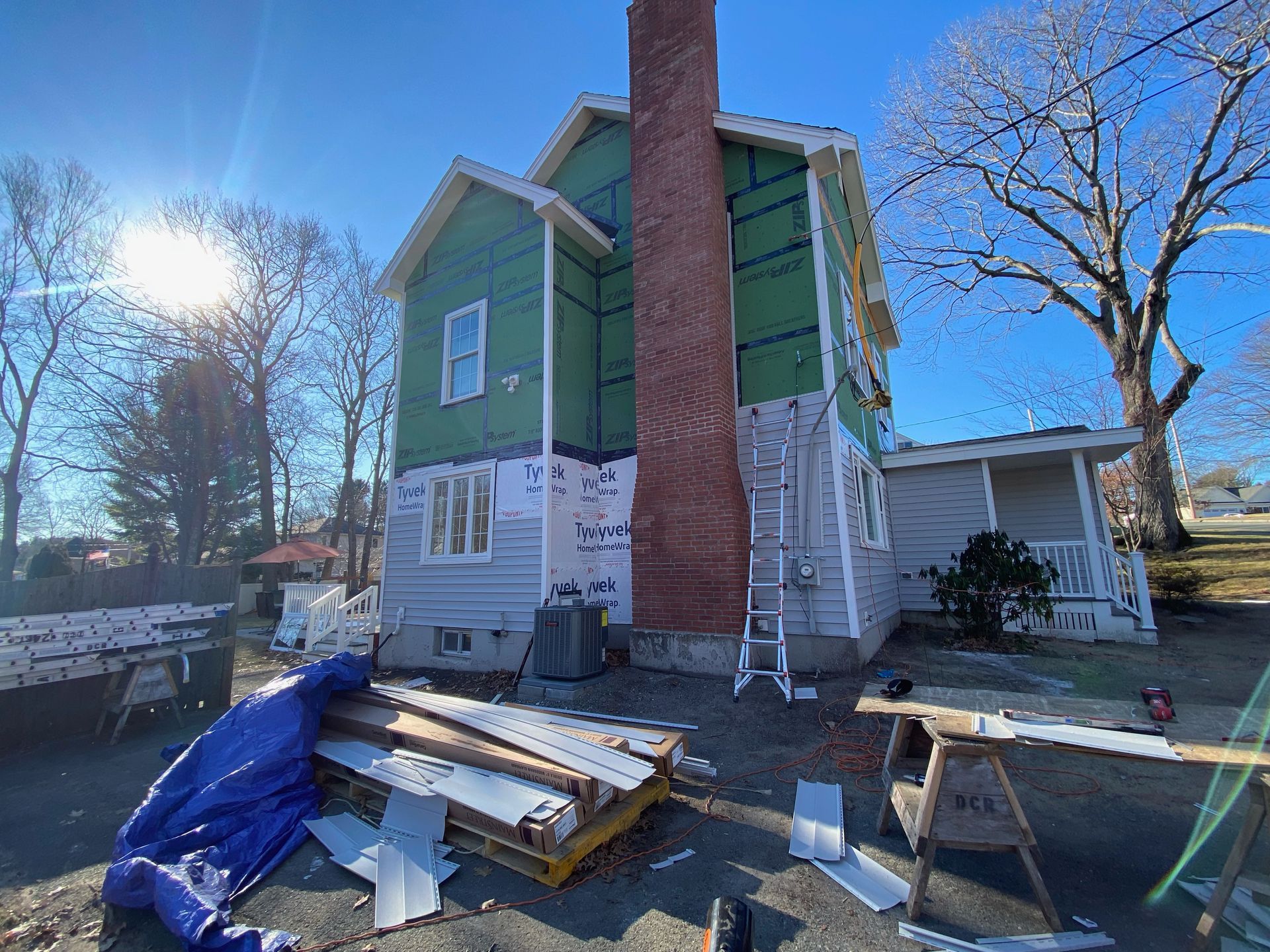House under construction; siding being installed. Green wrap, chimney, materials and tools on ground.