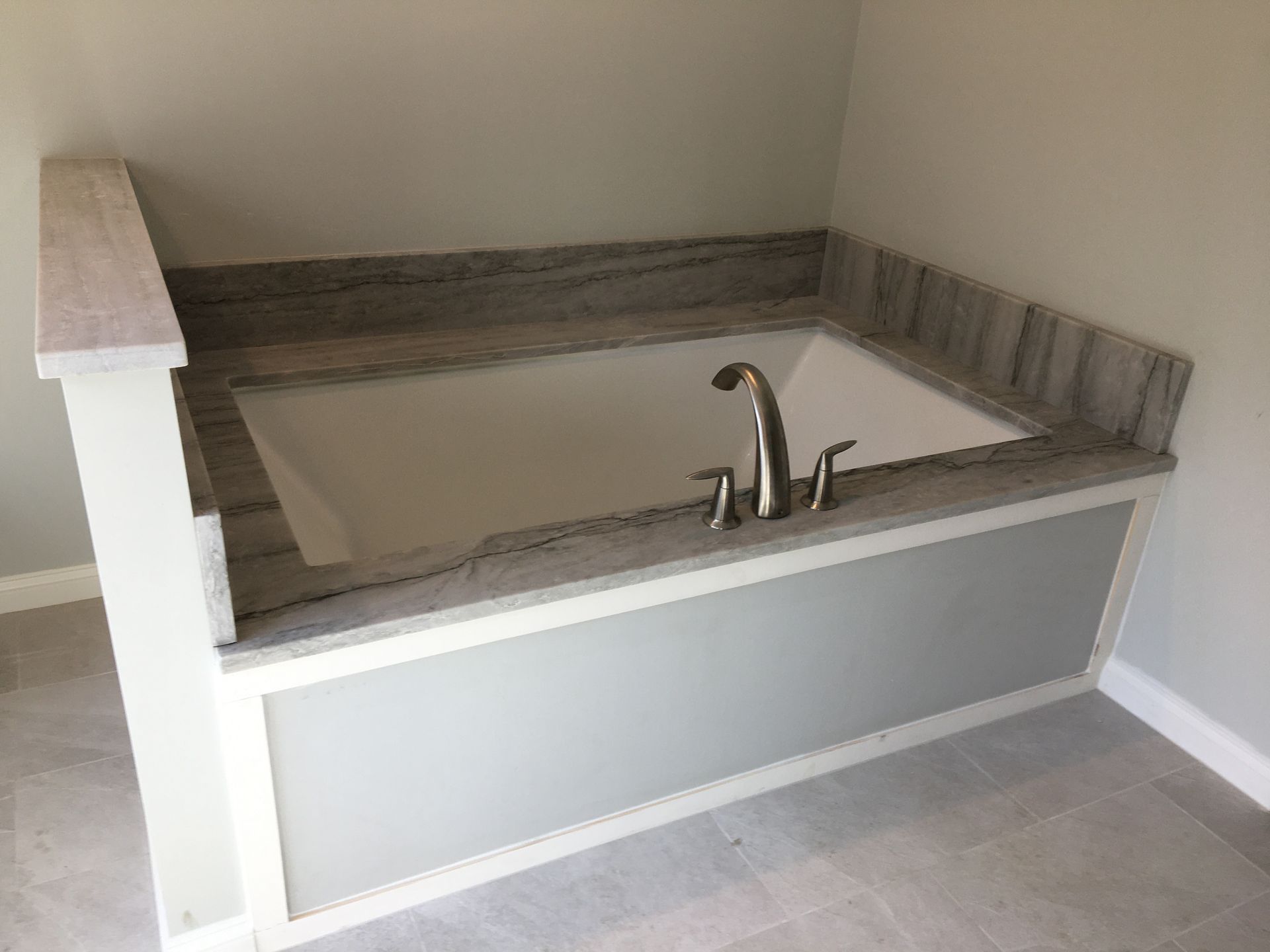 Built-in bathtub with grey countertop and faucet. Light grey wall and white framed base.