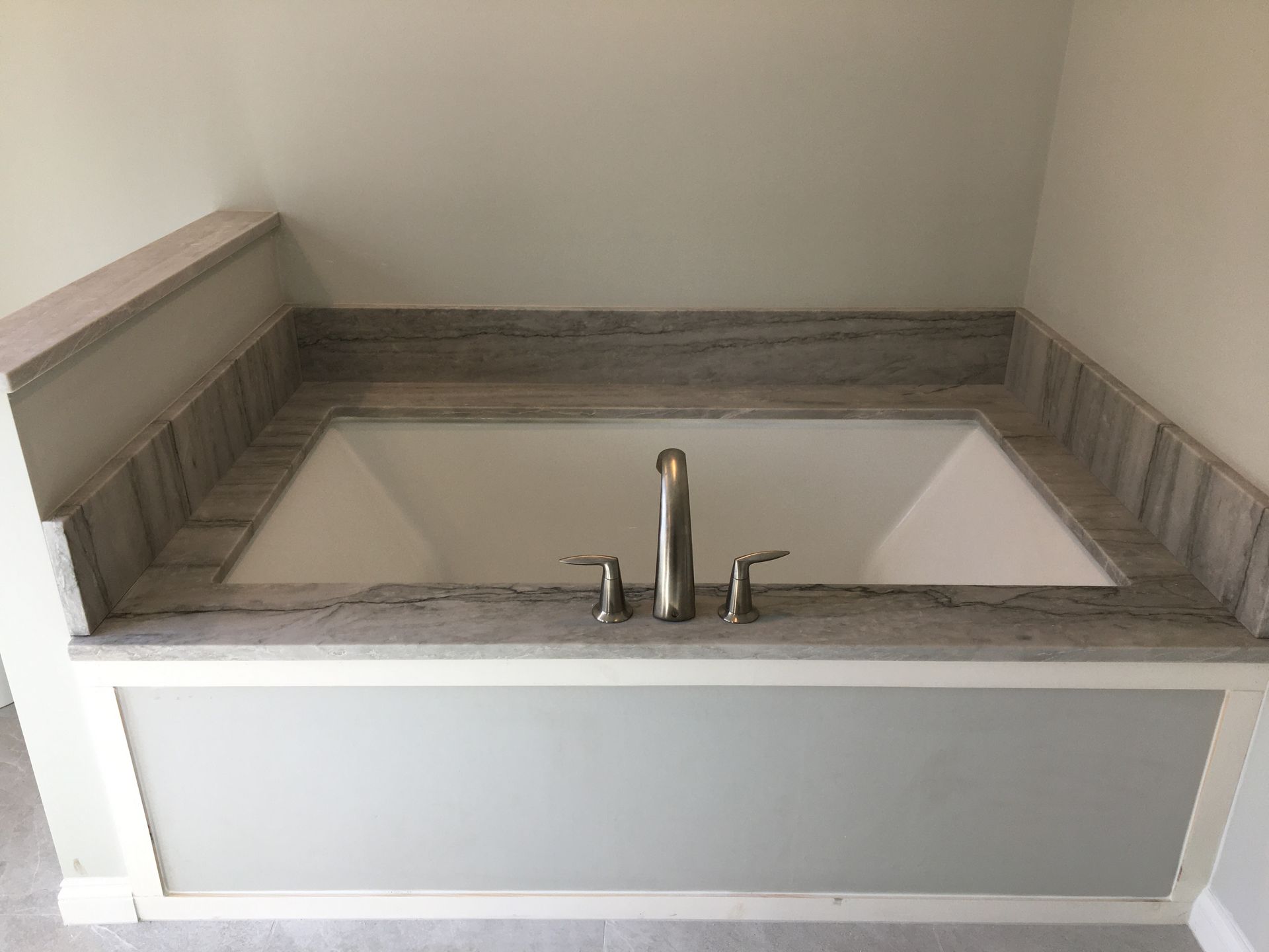 Built-in white bathtub with gray marble trim and silver faucet against a gray painted wall.