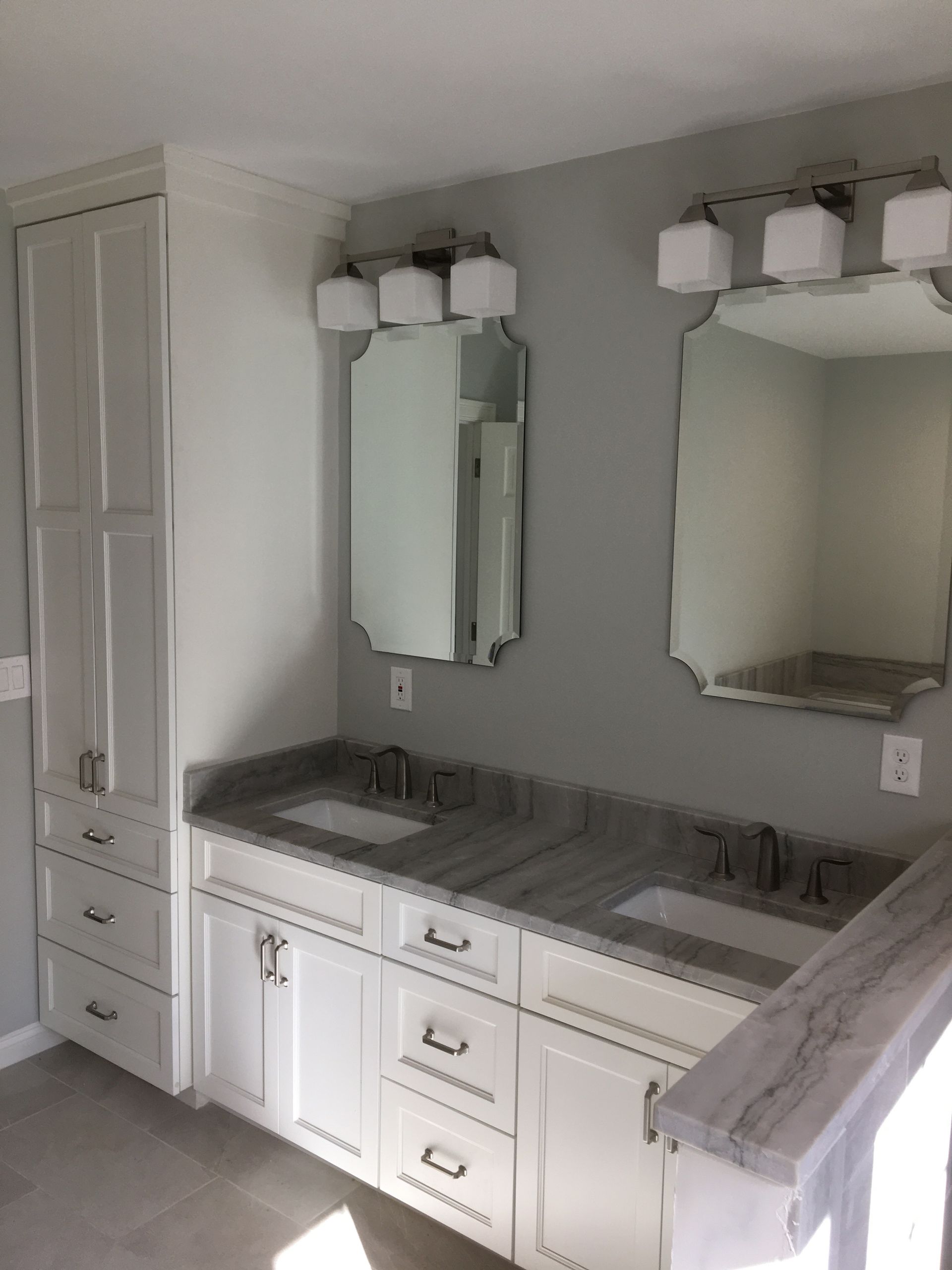 Bathroom with white vanity, gray countertop, mirrors, and light fixtures; gray walls and tall storage cabinet.