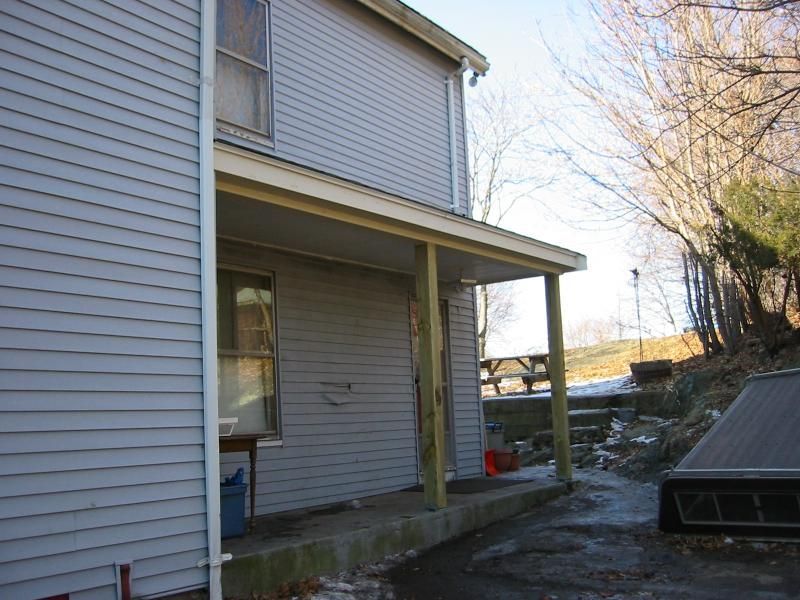 Side view of a two-story house with a porch. Gray siding, wooden supports, and a concrete walkway.