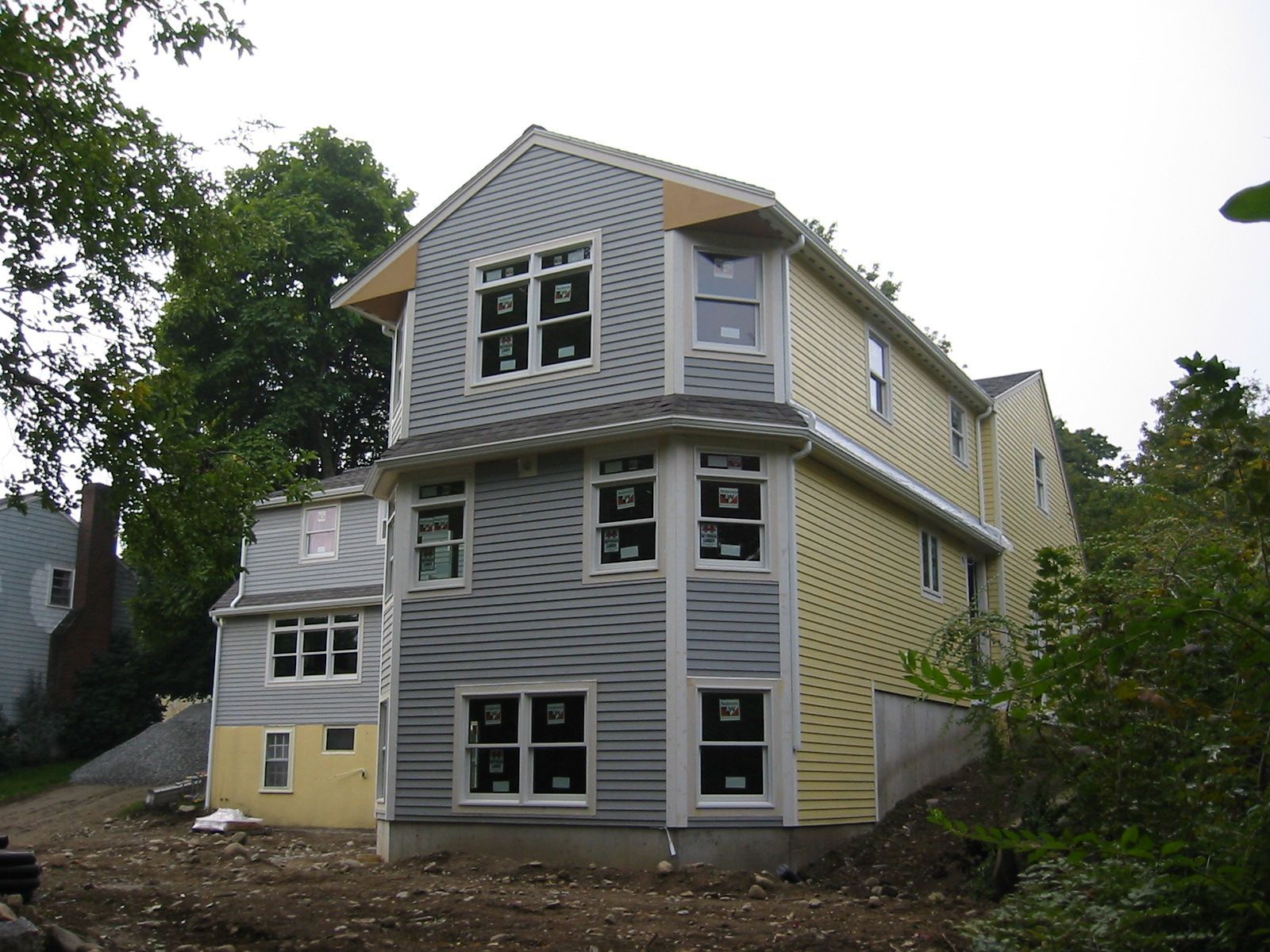 Two-story house under construction, yellow and grey siding, many windows, wooded background.